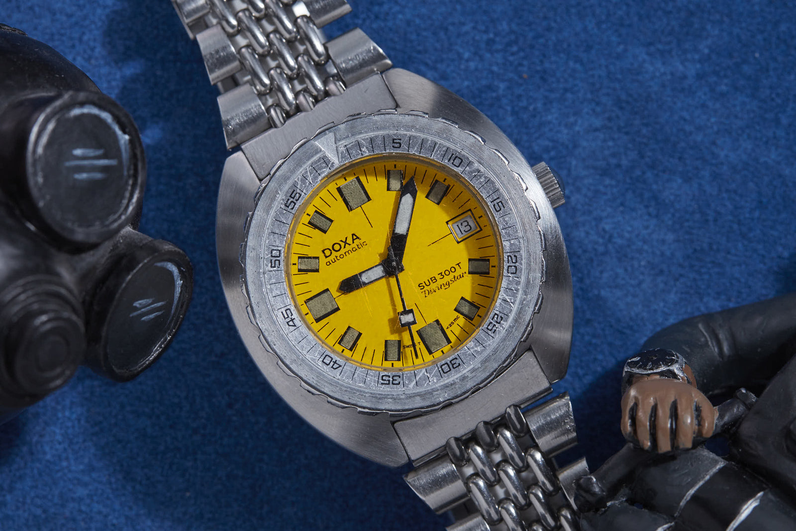 The DOXA Sub 300T Diving Star, a stainless steel dive watch with a bright yellow dial and date display, rests on a blue surface beside a gas mask and figurine’s hand, capturing DOXA's adventurous heritage.