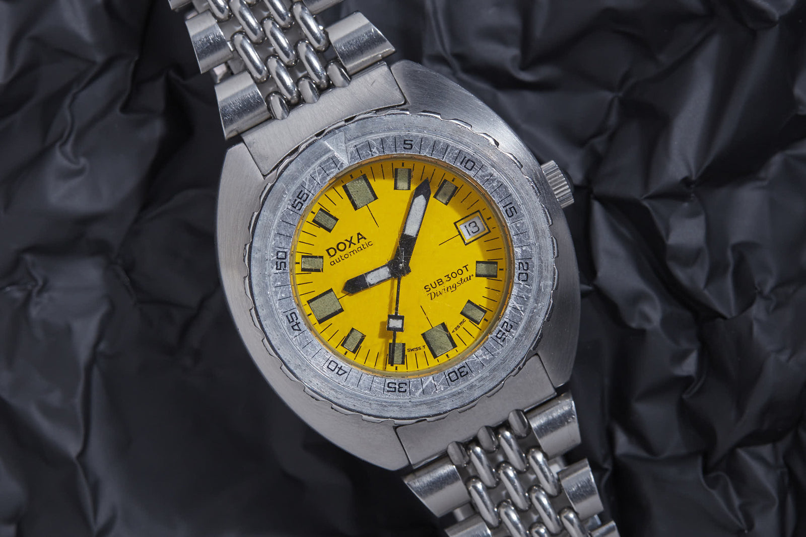 The DOXA Sub 300T Diving Star dive watch by DOXA features a stainless steel case, a bright yellow dial, black and orange hands, and a date window, shown on a crumpled black background.