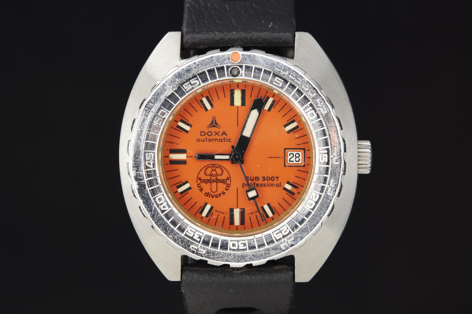 The DOXA Sub 300T Professional is a vintage diver’s watch featuring an orange dial, black hour markers, a date window at 3 o’clock, and a black leather strap, shown against a black background.