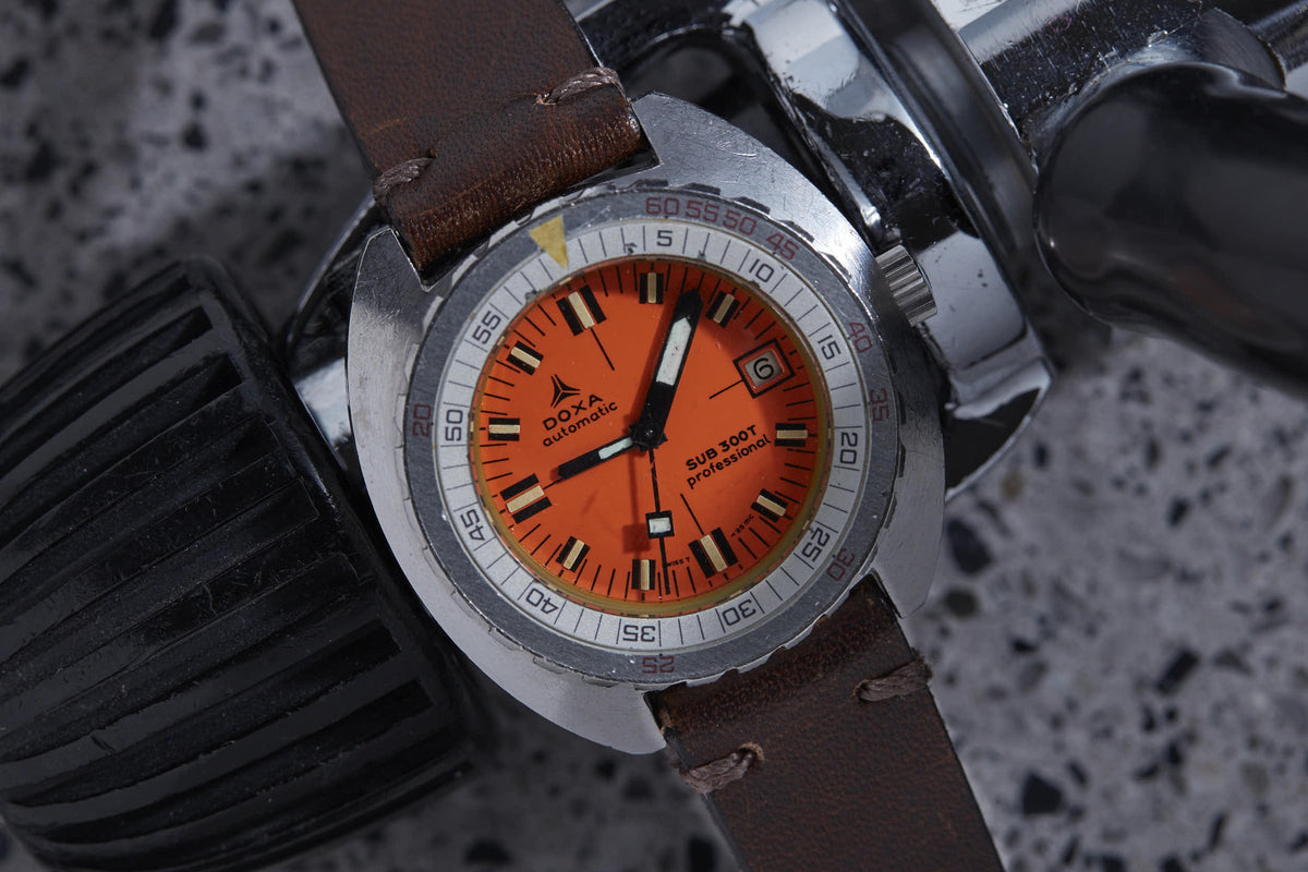 Stainless steel dive watch with bright orange dial, brown leather strap, and rotating bezel on textured surface.