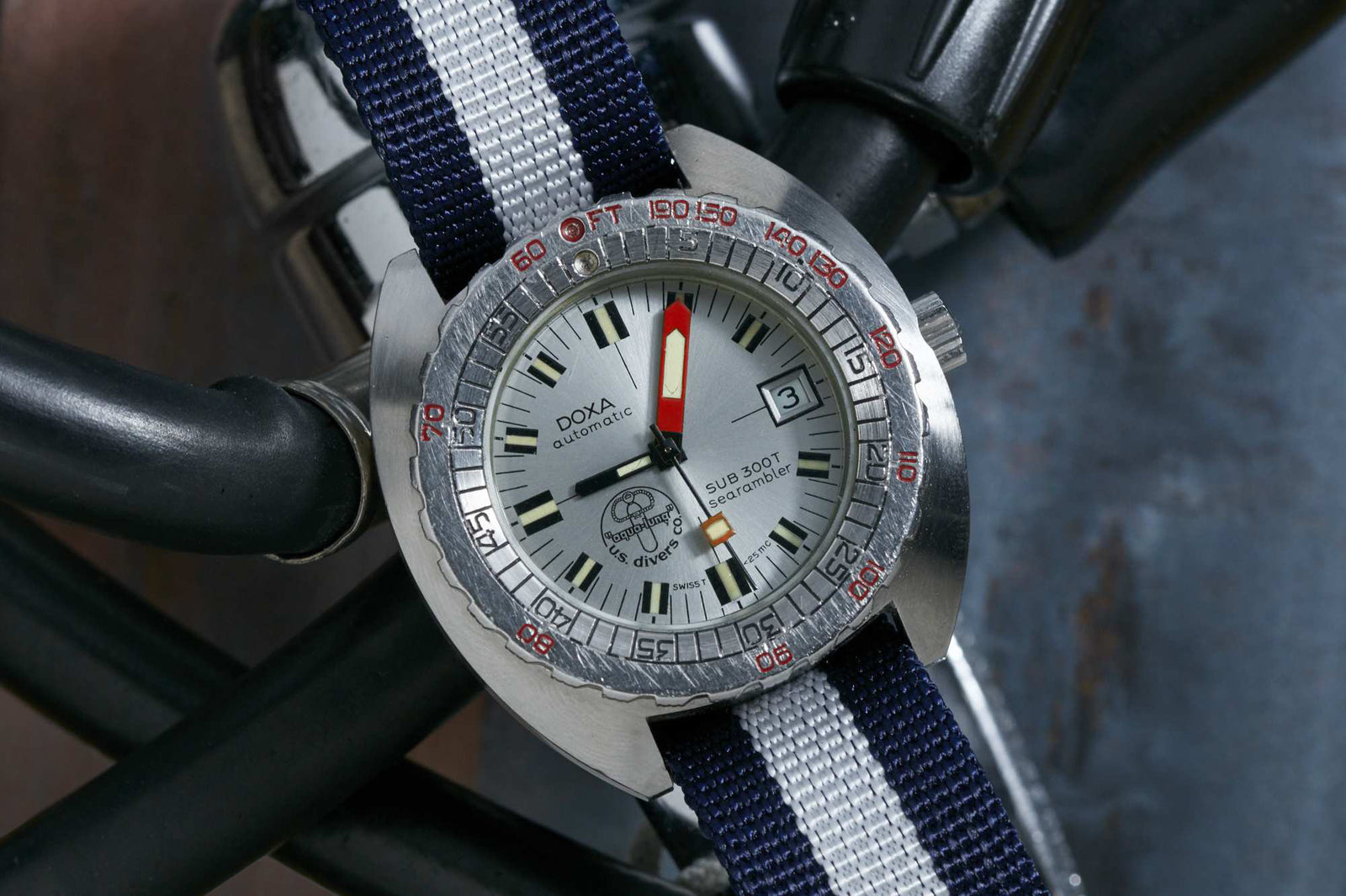 A DOXA Sub 300T Searambler dive watch with a blue and white striped fabric strap rests on black cables, featuring a silver case, white dial, orange minute hand, date at 3 o’clock, and red bezel markings.