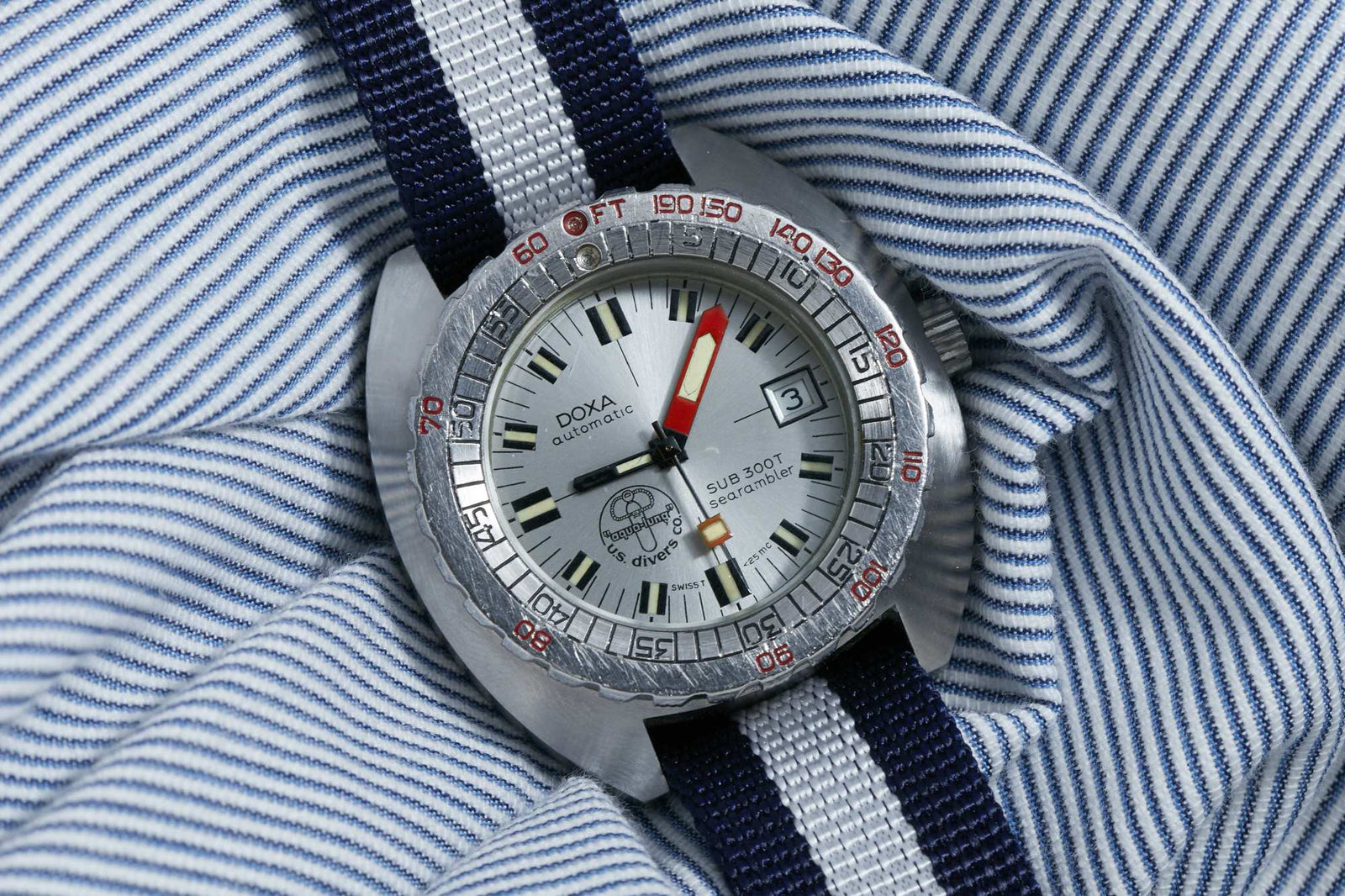 A DOXA Sub 300T Searambler dive watch with a white dial, date display, red minute hand, and a black/navy/gray striped nylon strap rests on blue and white striped fabric.