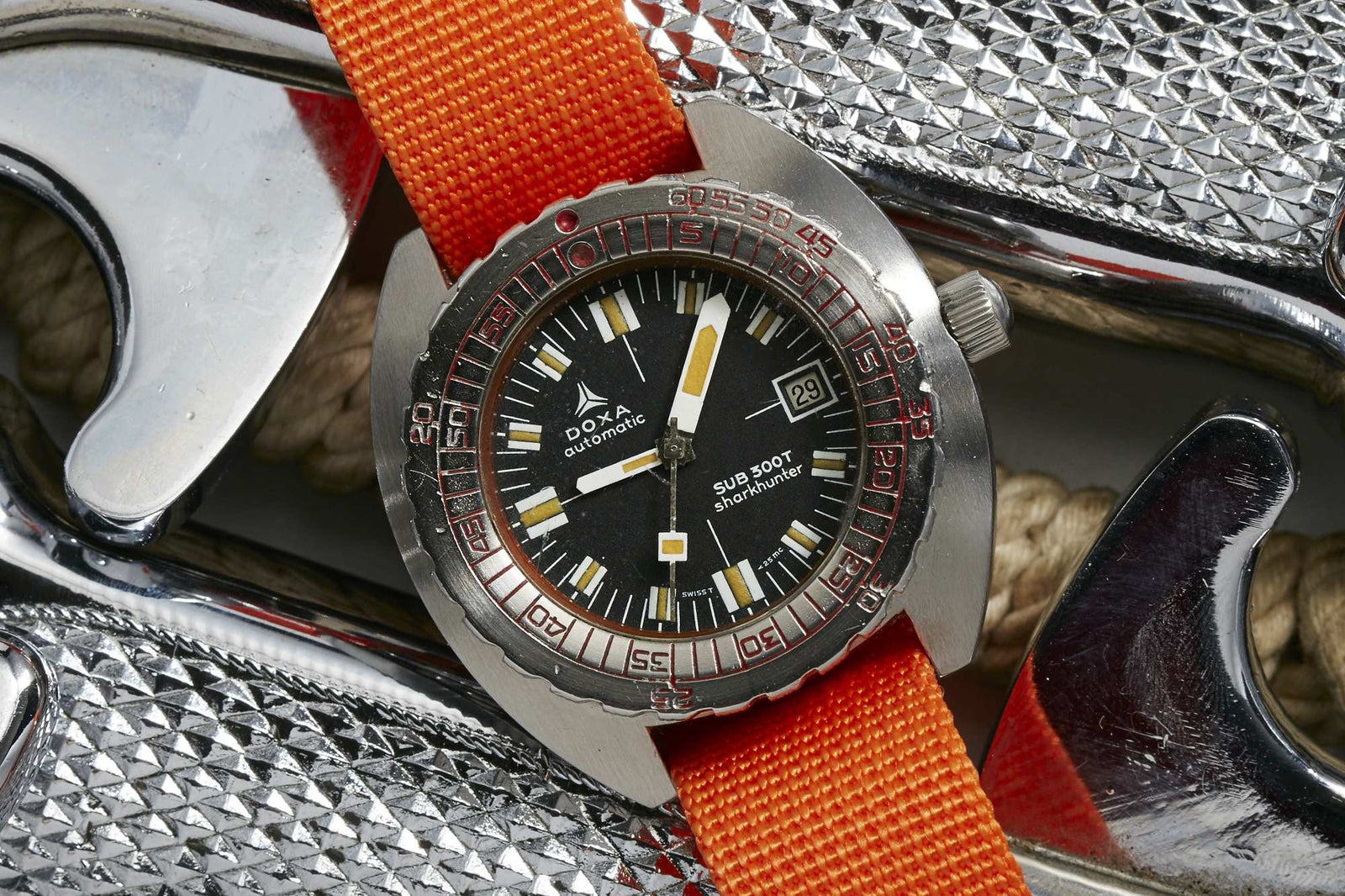 A DOXA Sub 300T Sharkhunter dive watch with an orange strap lies on silver textured metal objects, a rope in the background. The black dial sports orange and white accents plus a date display, showcasing DOXA’s renowned craftsmanship.