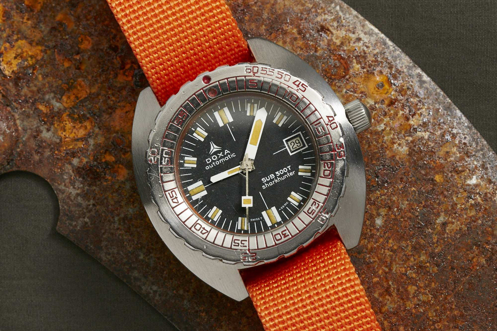 A DOXA Sub 300T Sharkhunter dive watch with a black dial, bold white and orange markers, date window at 3 o’clock, and an orange strap rests on a rusty metal surface—a true icon for dive watch enthusiasts.