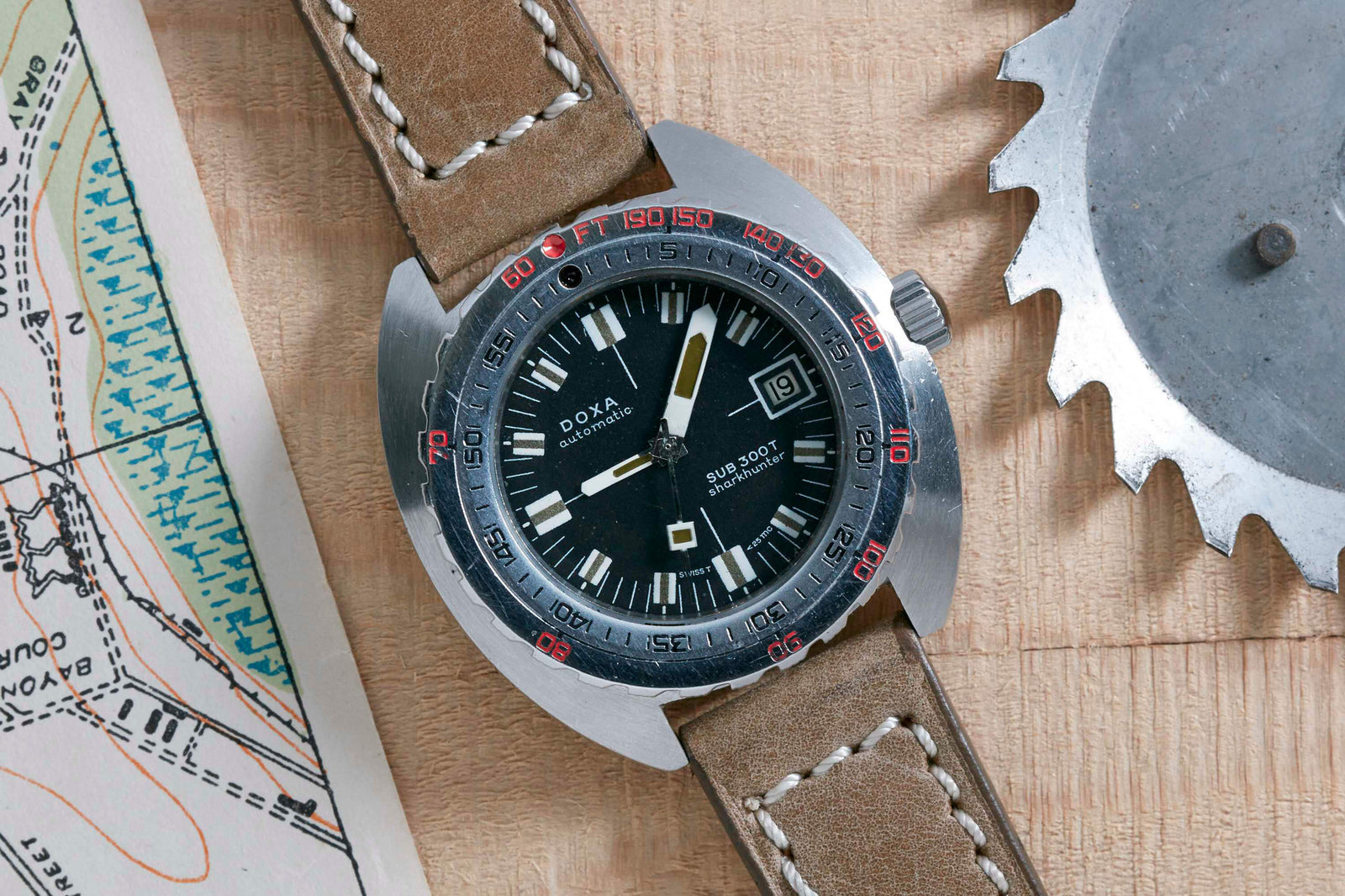 A DOXA Sub300T Sharkhunter dive watch with a brown leather strap sits on a wooden surface beside a map and metal gear. Its black dial features a date display, while red numerals accent the bezel.