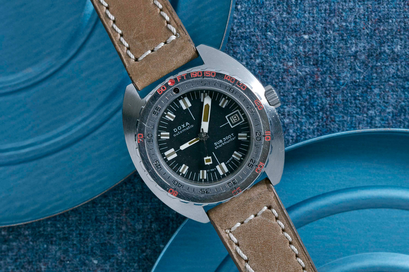 A DOXA Sub300T Sharkhunter diving watch with a brown leather strap, black dial, and silver bezel with red numbers, shown on a blue textured surface alongside blue plates.