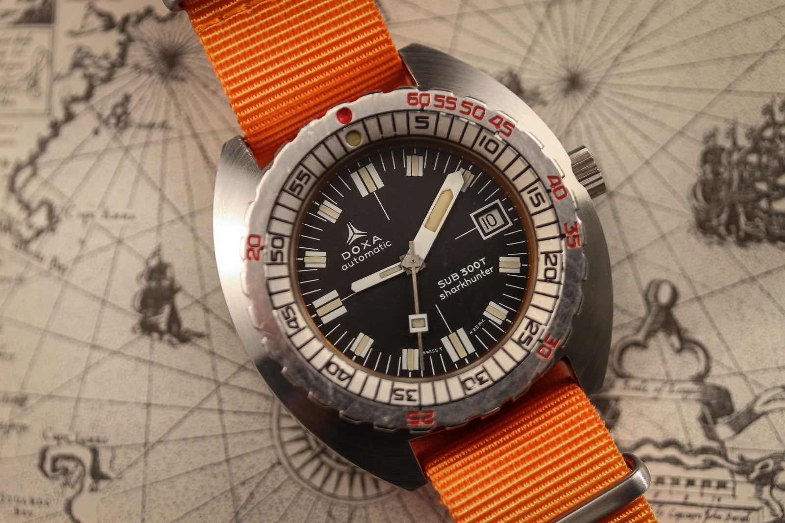 A DOXA Sub 300T Sharkhunter diver's watch with a black dial, orange minute hand, and orange fabric strap rests on a nautical map. Its date display and bold white bezel indices enhance its classic underwater style.