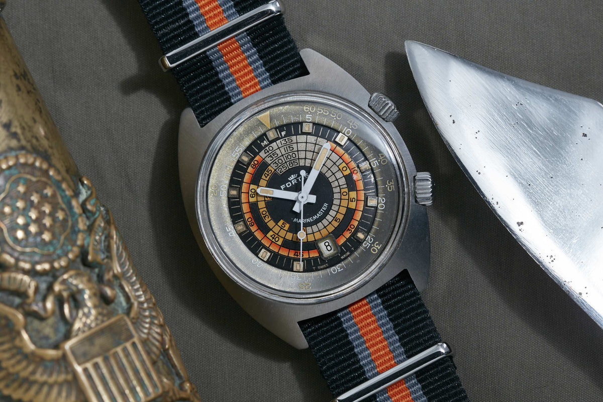 A Fortis Marinemaster automatic wristwatch with a black, orange, and silver dial and a matching strap lies on a dark surface next to a metallic object and a carved brass item.