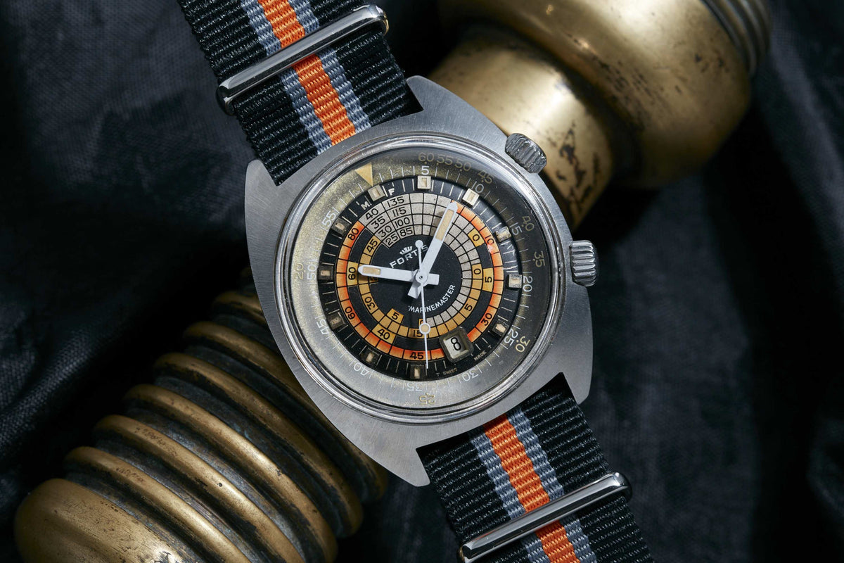 The Fortis Marinemaster by Fortis, an automatic wristwatch featuring a complex black, orange, and cream dial, black case, and striped nylon strap, rests on brass objects against a dark background.
