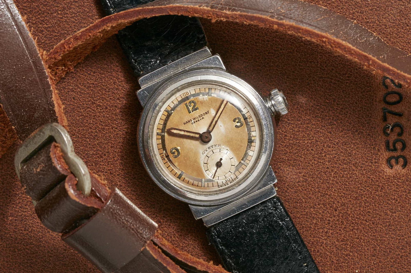 The Hans Wilsdorf Oyster by Hans Wilsdorf features a vintage look with a worn leather strap, beige and silver dial, and black numerals. Displayed on brown leather marked 353702. Detailed description coming soon.