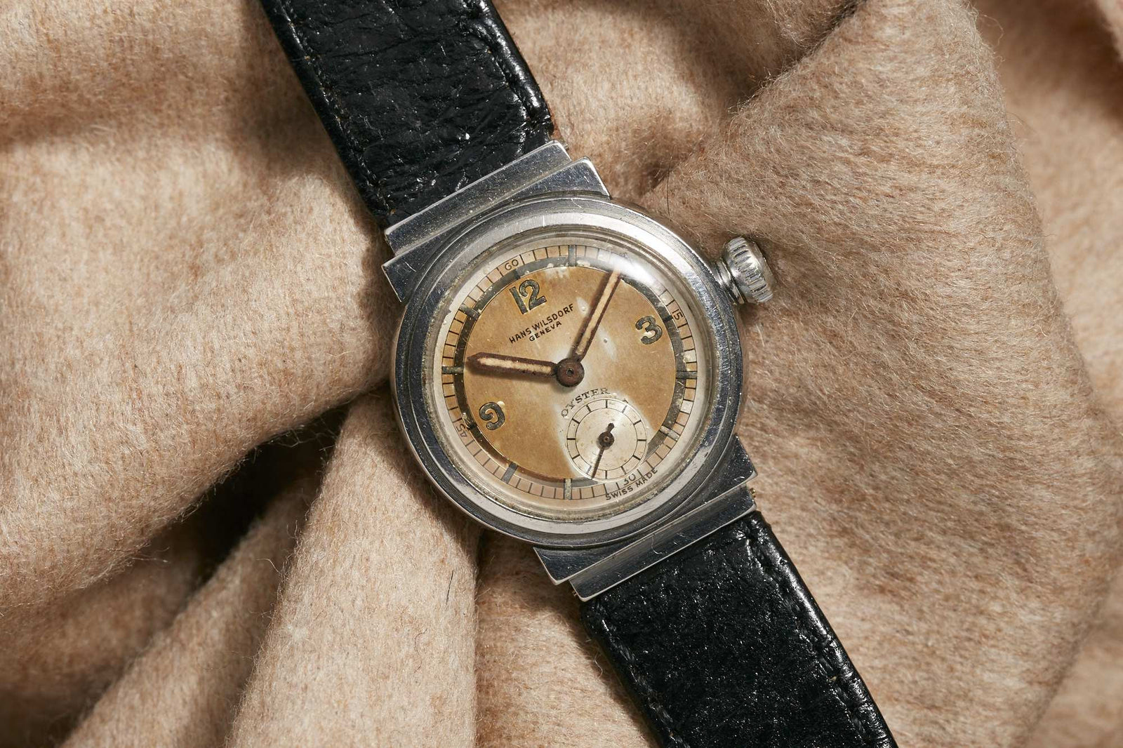 The Hans Wilsdorf Oyster by Hans Wilsdorf features a vintage round dial with Arabic numerals, metal case, and black leather strap shown on soft beige wool. Detailed description coming soon.