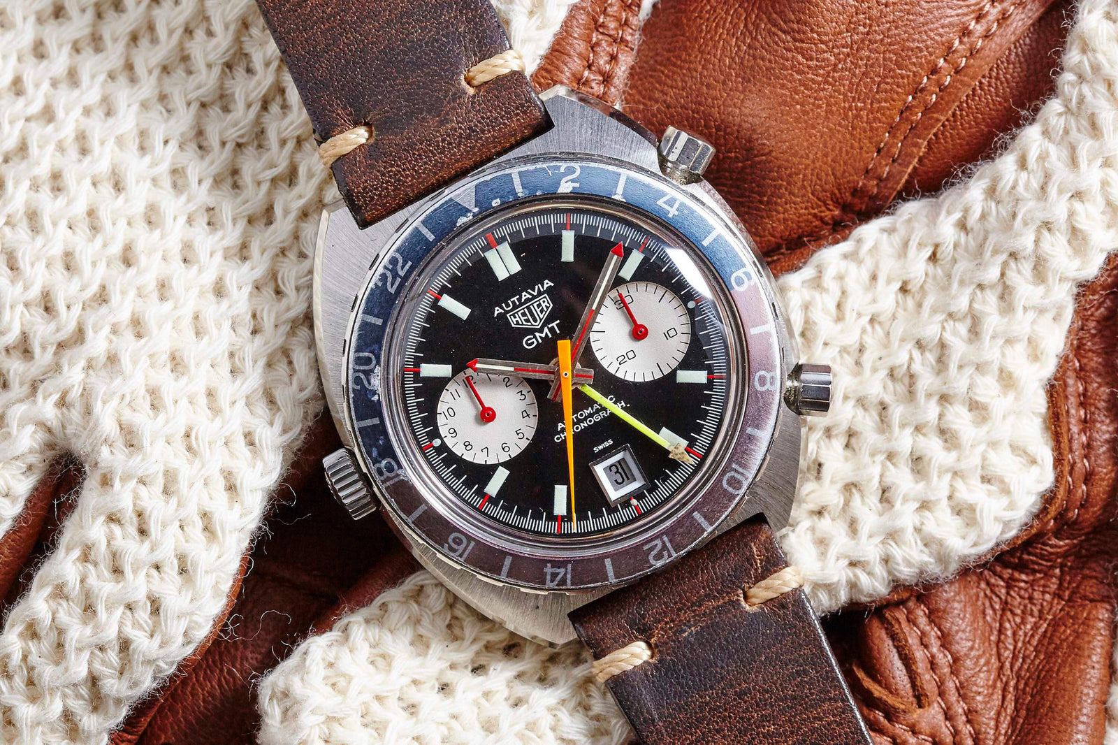 A Heuer GMT 1163 chronograph with a brown leather strap rests on cream knit gloves and brown leather, featuring a black dial, red accents, and a blue-red GMT bezel. Description coming soon.
