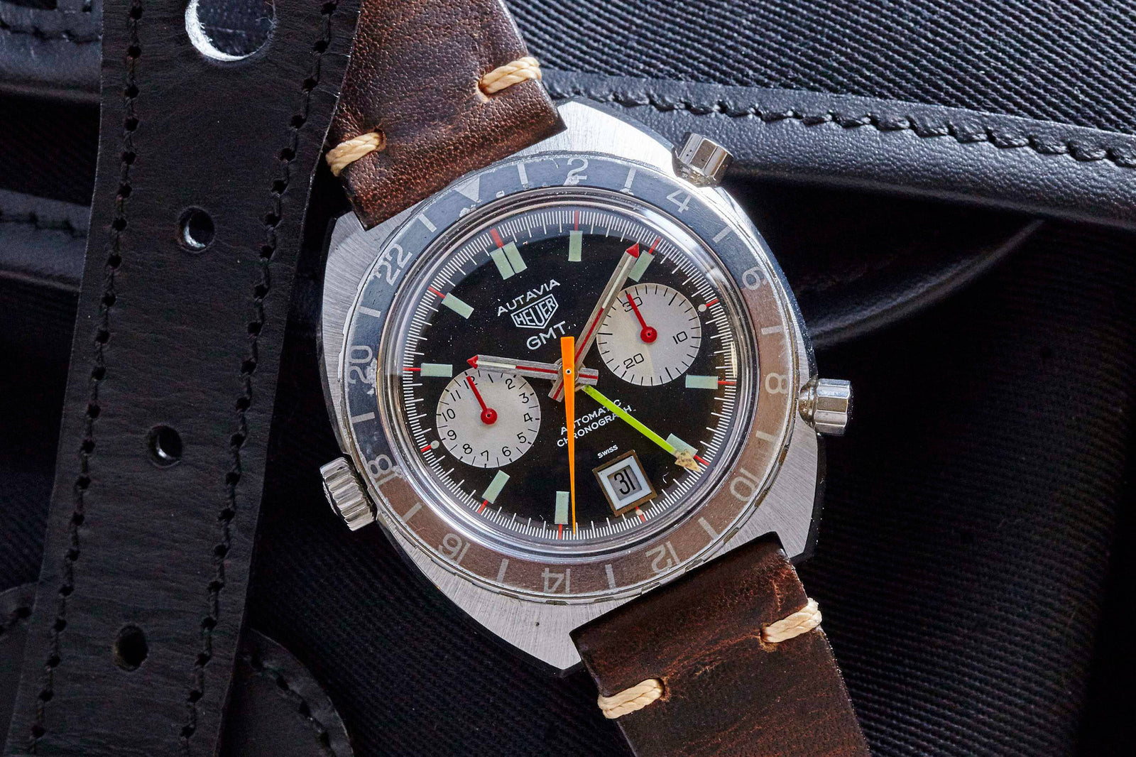 A Heuer GMT 1163 watch with a black dial, two subdials, red and yellow hands, date window at 6 o’clock, and leather strap rests on black leather. Description coming soon.