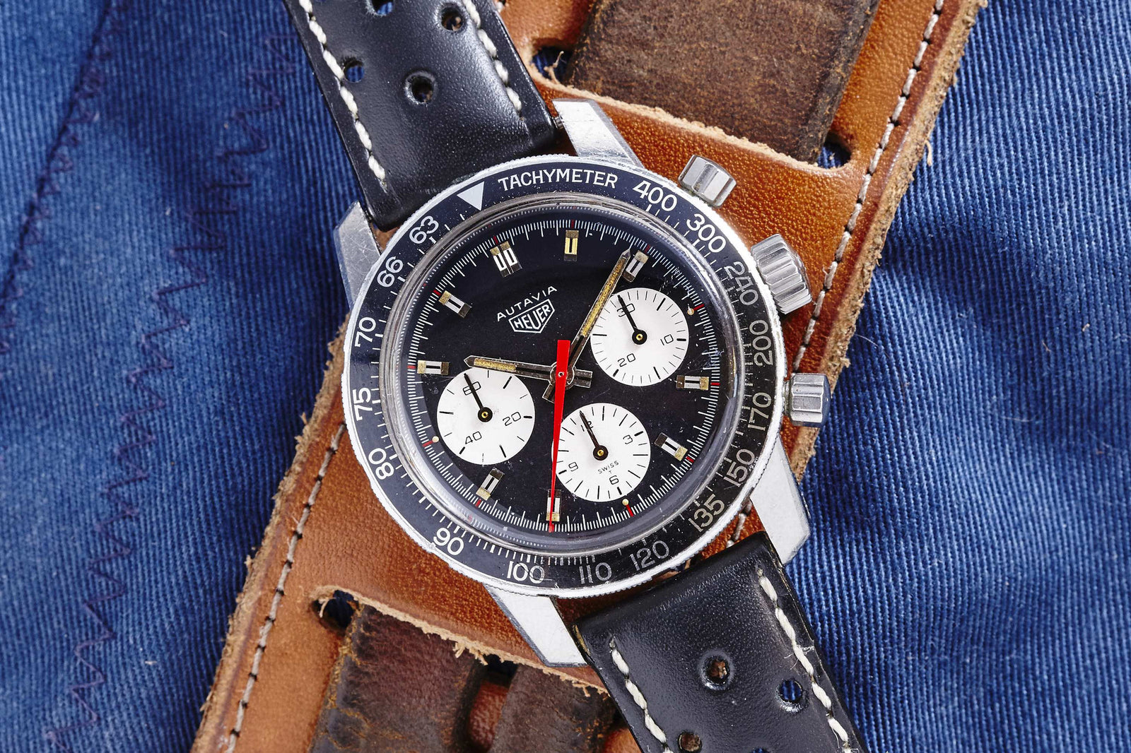 A vintage Heuer Autavia by Heuer, featuring a black bezel, black dial, three white subdials, and a brown leather strap, rests on blue denim—a classic manually-wound chronograph ideal for collectors.