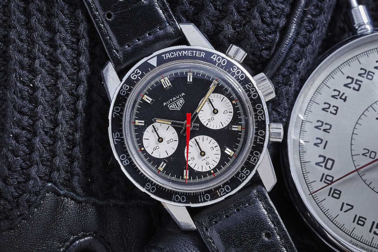 A close-up of the Heuer Autavia by Heuer, a motorsports classic with a black dial, three white subdials, red seconds hand, black leather strap, and tachymeter bezel on a knitted black surface beside a stopwatch.