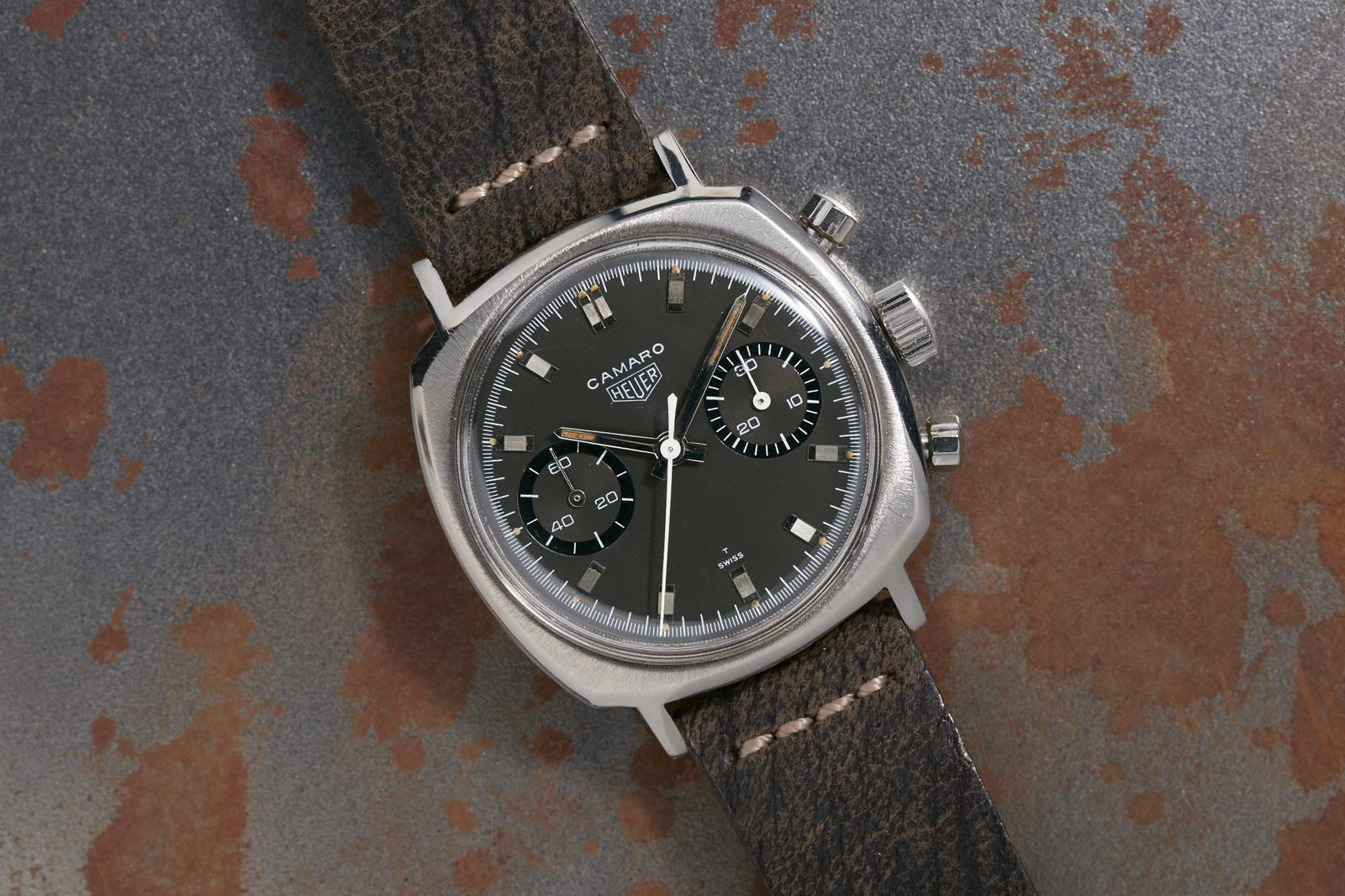 A vintage Heuer Camaro chronograph with a black dial, two subdials, silver markers, and a worn brown leather strap lies on a textured, rusted metal surface.