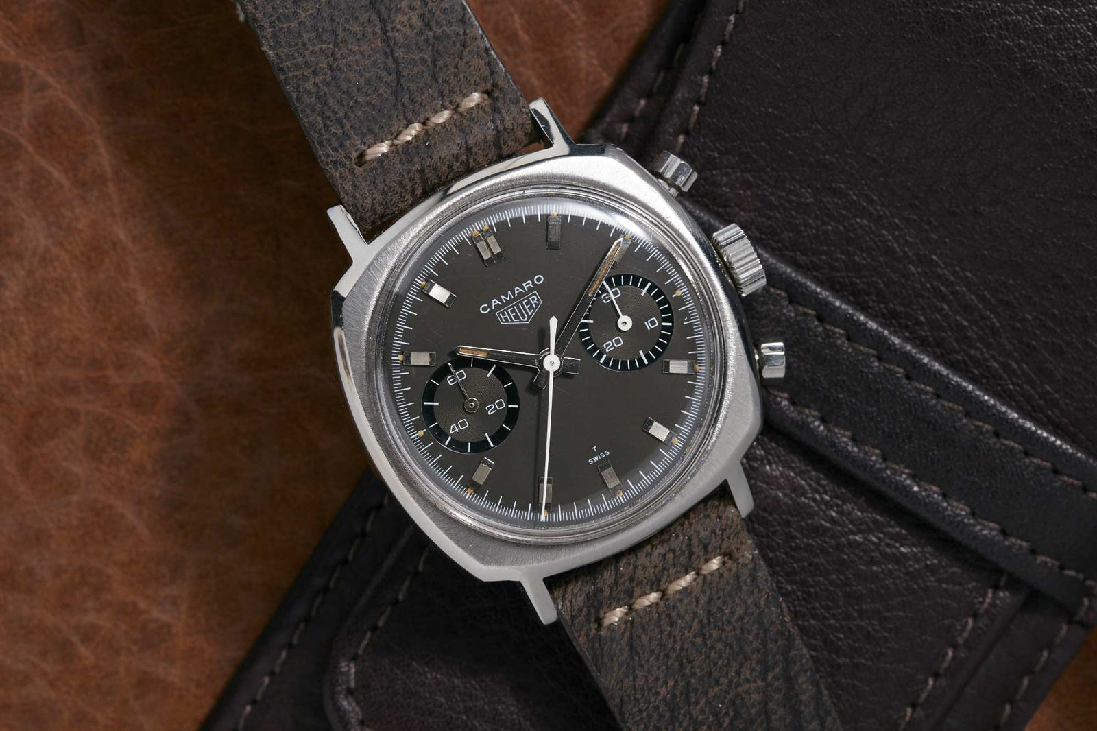 A Heuer Camaro chronograph by Heuer, featuring a grey leather strap and black dial with two sub-dials, rests on a dark leather surface accented with brown in the background.