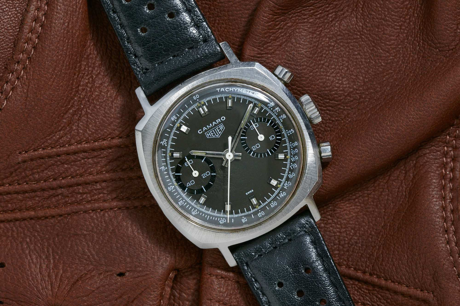 A Heuer Camaro 7743N chronograph with black dial, tachymeter scale, and two subdials on a black leather strap is shown atop a brown leather surface.