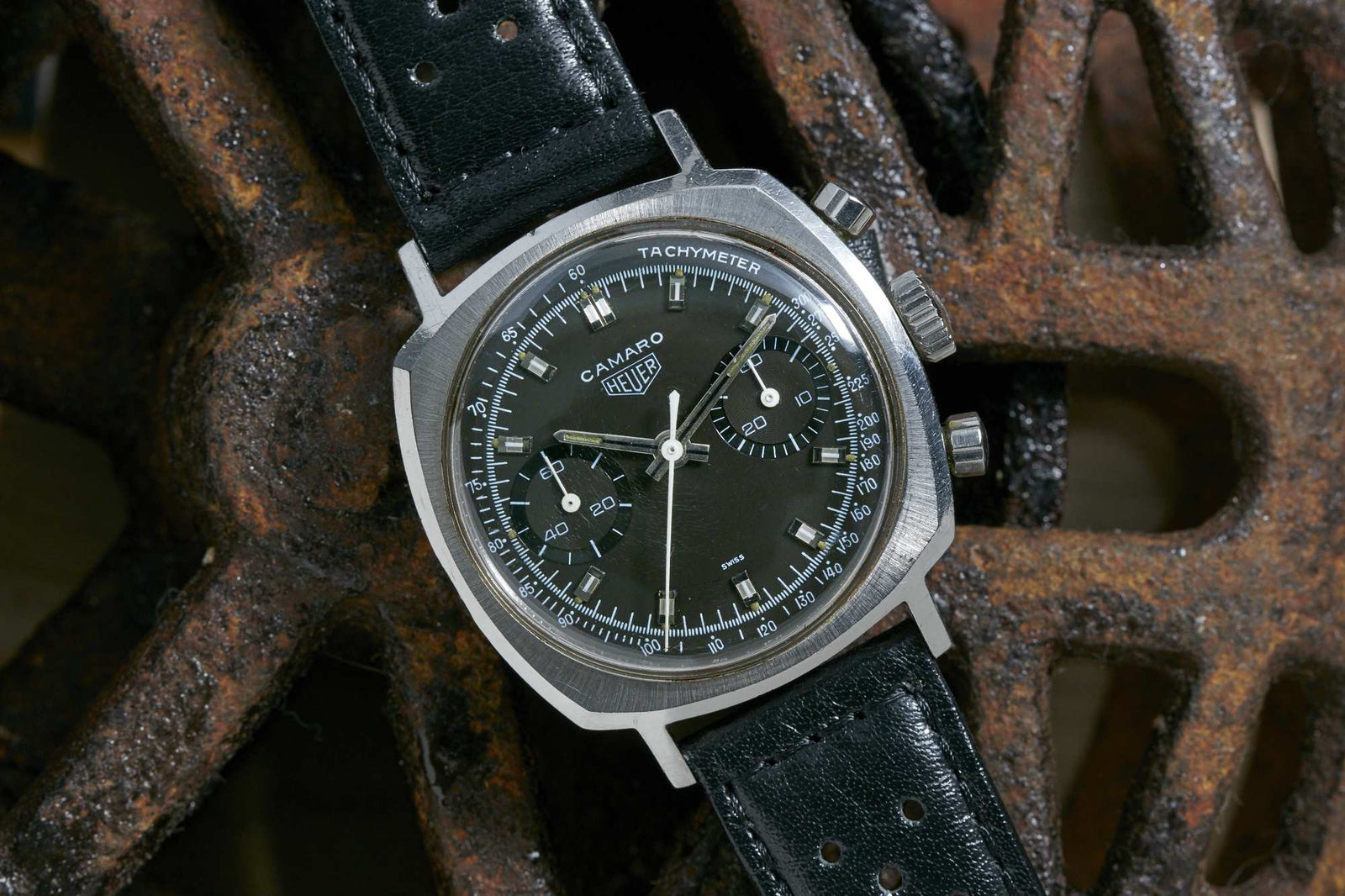 A Heuer Camaro 7743N chronograph wristwatch by Heuer, featuring a black dial and leather strap, is showcased against a rusty metal wheel backdrop.