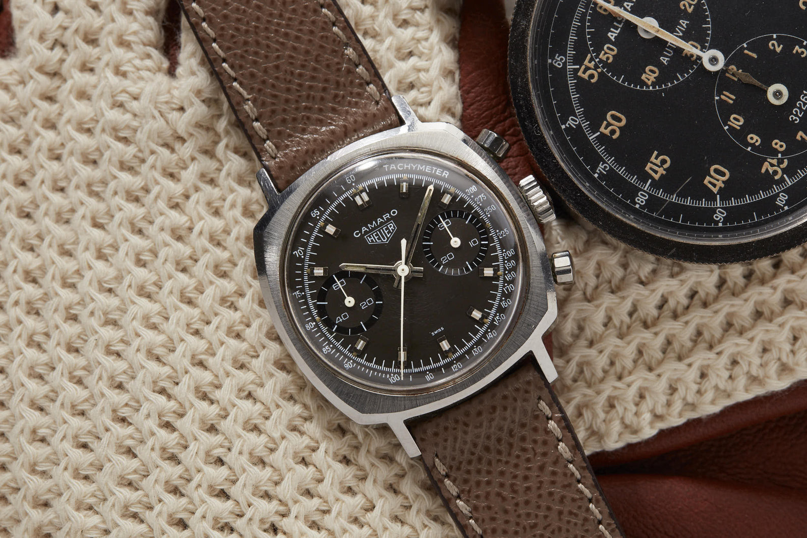 A close-up of a Heuer Camaro wristwatch by Heuer with a brown leather strap, shown on cream knitted fabric beside part of another black-dial vintage racing chronograph.