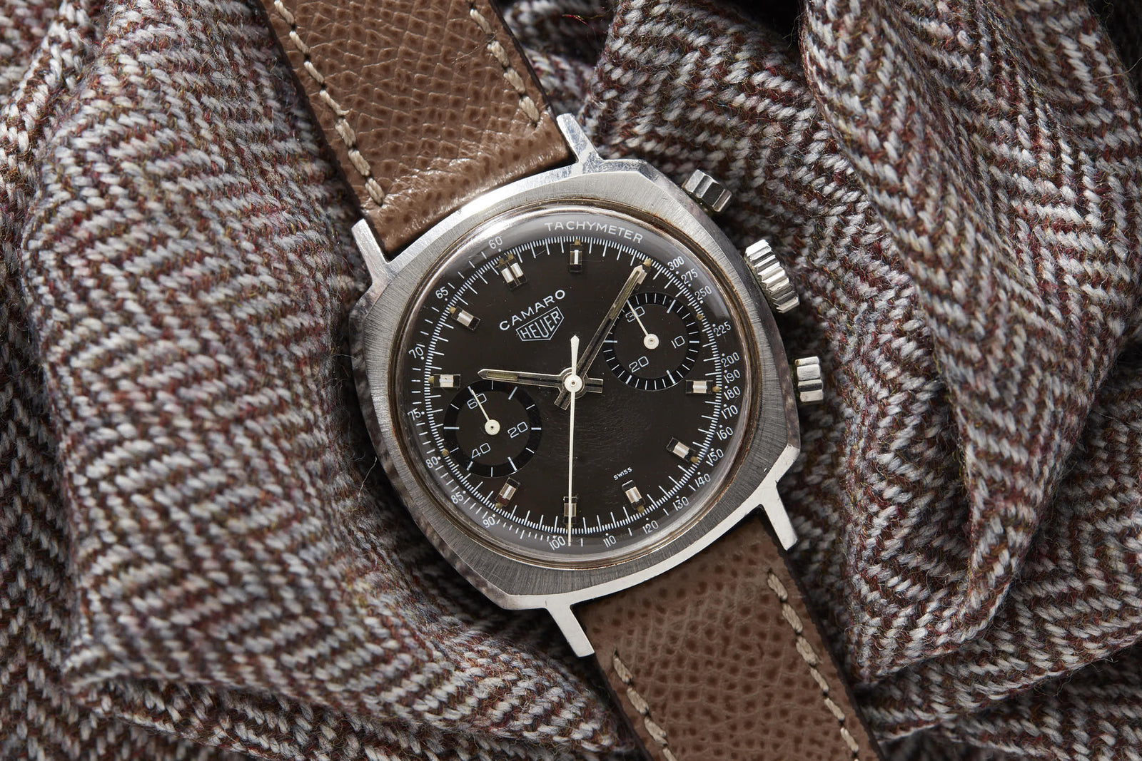 A vintage racing chronograph, the Heuer Camaro watch by Heuer with a brown leather strap rests on textured houndstooth fabric. Its black dial features silver markers and three subdials, evoking classic motorsport style.