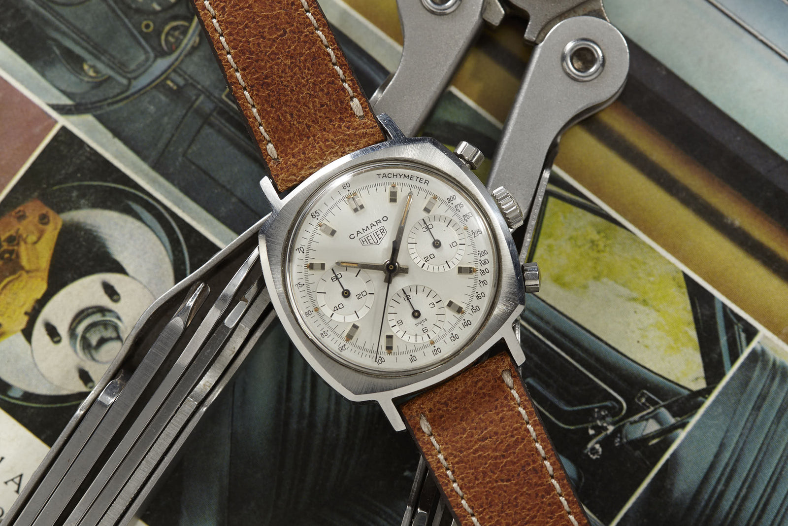 A Heuer Camaro vintage chronograph by Heuer with a brown leather strap rests on assorted tools and colorful illustrated papers.