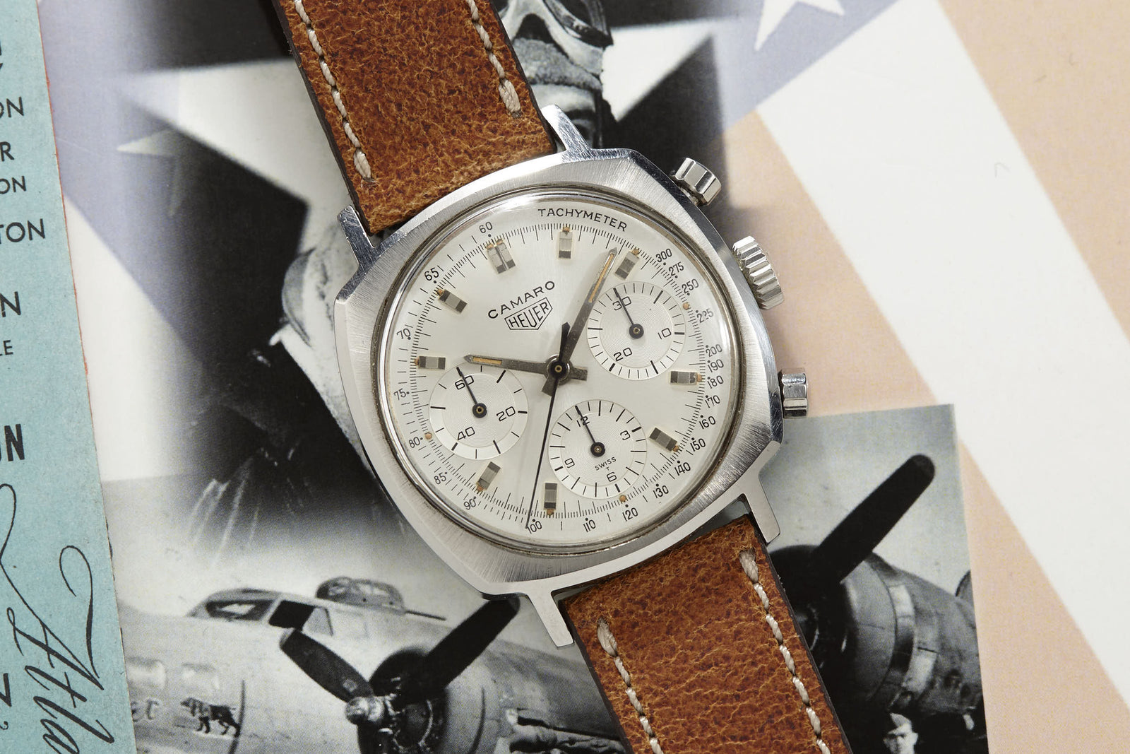 A vintage Heuer Camaro tachymeter chronograph wristwatch by Heuer with a brown leather strap rests on an aviation-themed surface. The watch features a silver dial, three subdials, and black markers.