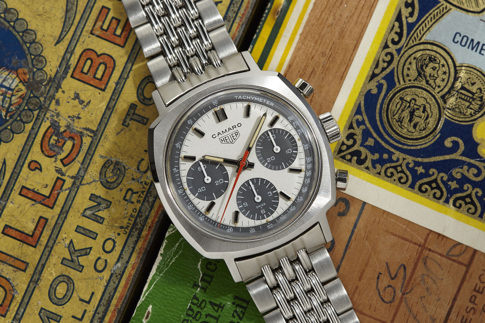 A Heuer Camaro Exotic Dial chronograph from Heuer with a classic panda dial is displayed on vibrant vintage matchbox labels and wooden surfaces.