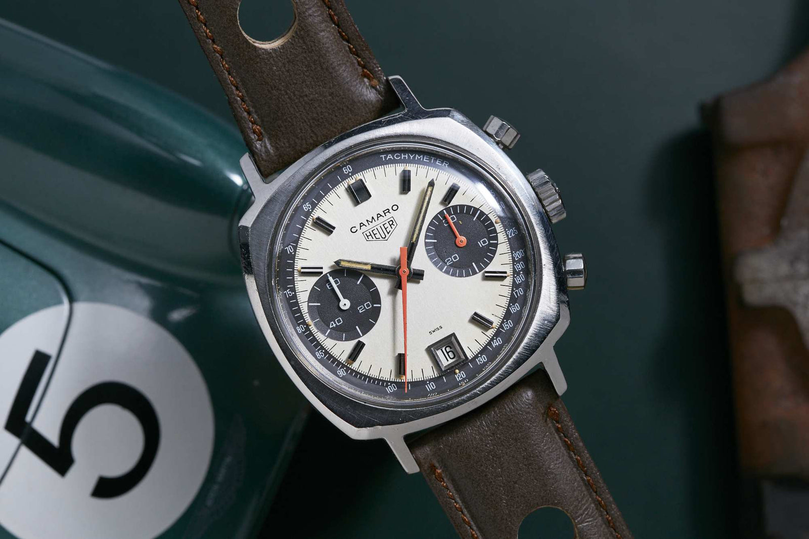 The Heuer Camaro Date Exotic Dial by Heuer features a beige dial, black subdials, brown leather strap, date display, and tachymeter scale next to a green object marked with a white circle and the number 5.