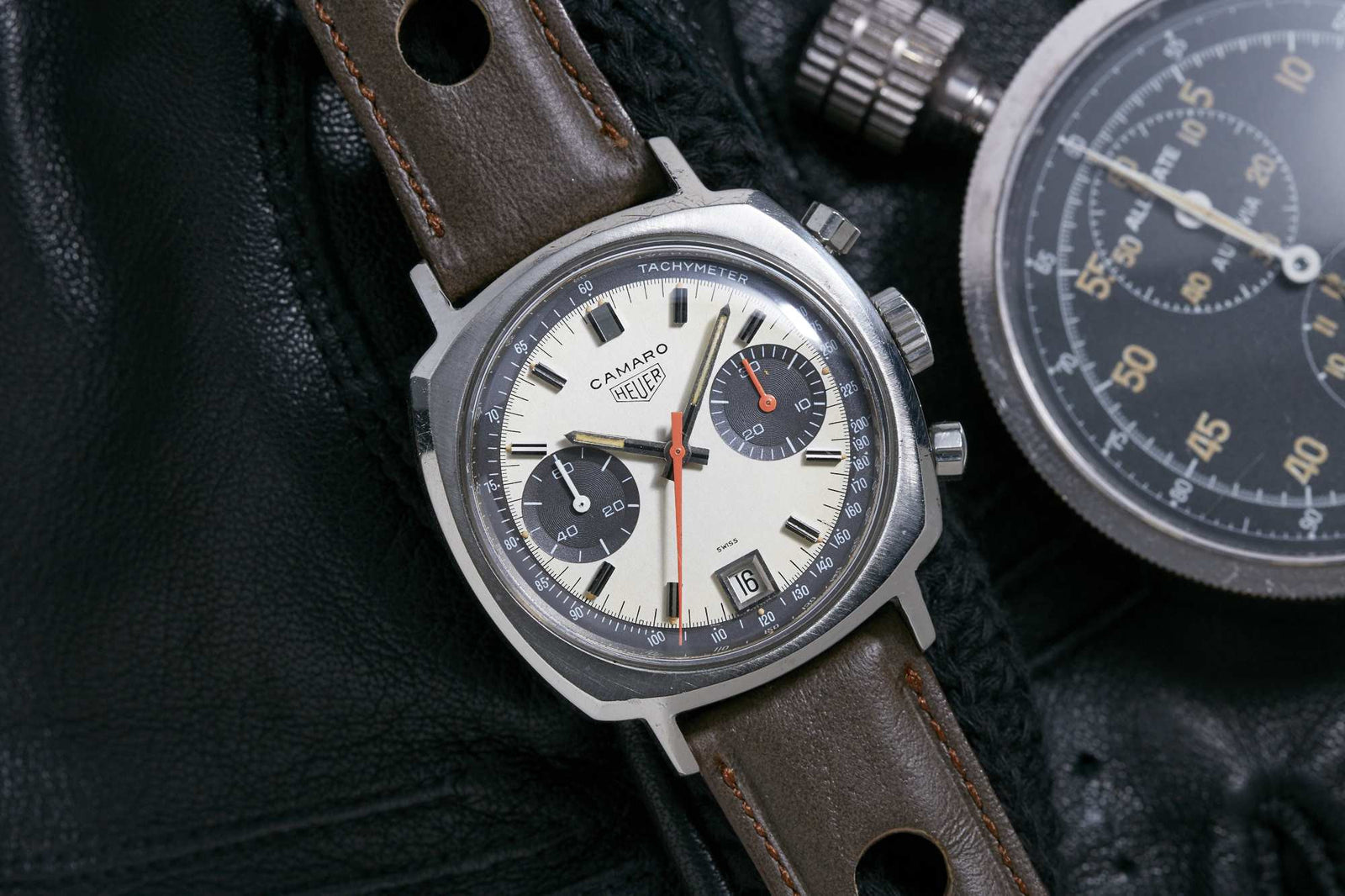 A Heuer Camaro Date Exotic Dial watch by Heuer, featuring a cream and black dial, tachymeter scale, brown leather strap, and red chronograph hand, rests on black leather beside a silver stopwatch.