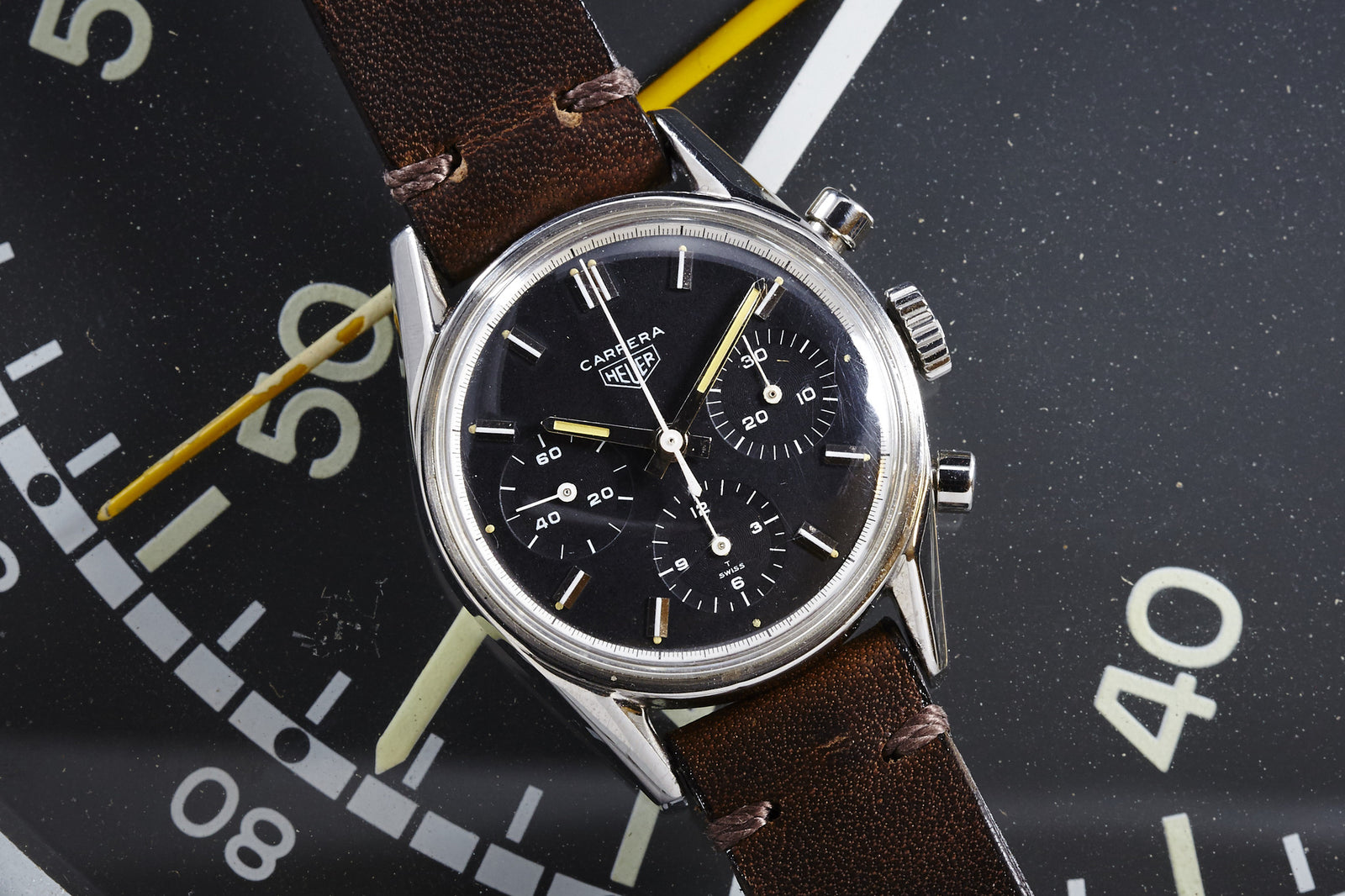 A vintage Heuer Carrera 2447N chronograph with a black dial, three subdials, and a brown leather strap rests atop a large clock face showing 40, 50, and 60.