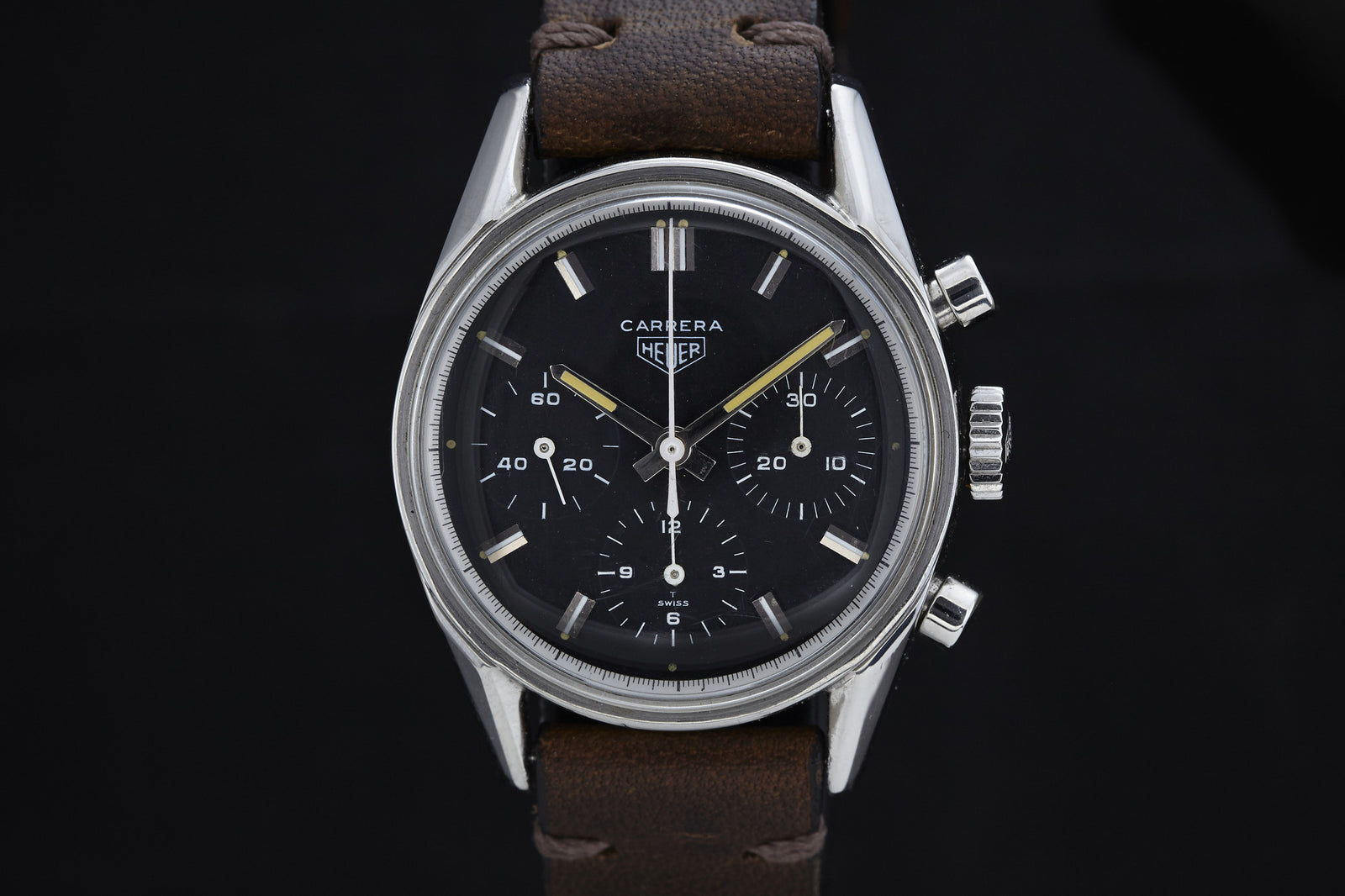 A close-up of a Heuer Carrera 2447N wristwatch features a Valjoux 72 chronograph, black dial, three subdials, silver markers, and a brown leather strap set against a black background.