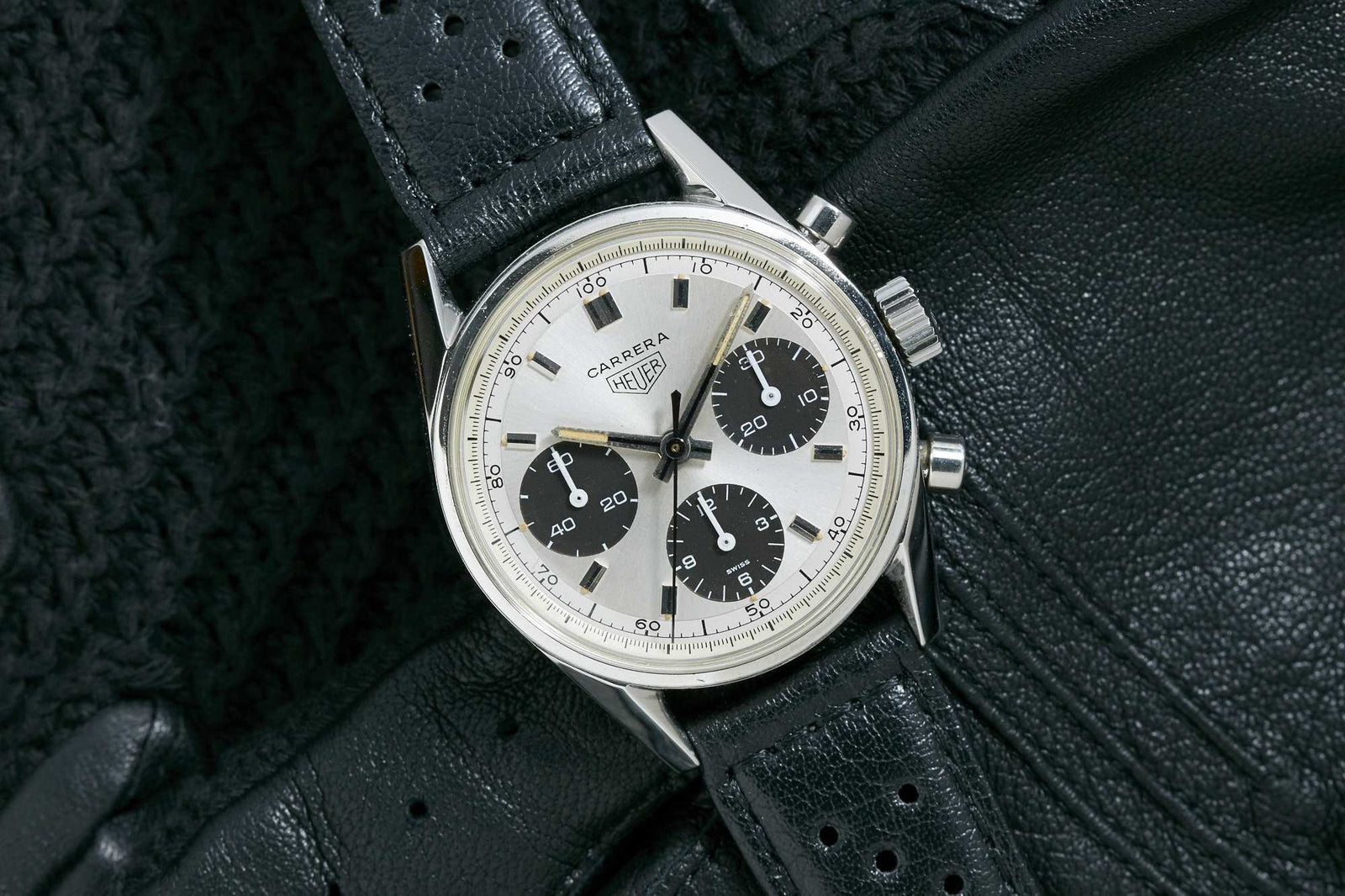 A vintage Heuer Carrera Ref. 2447SND chronograph with a silver dial and three black subdials is shown on a perforated leather strap, resting on black gloves and a textured surface. Detailed description coming soon!.