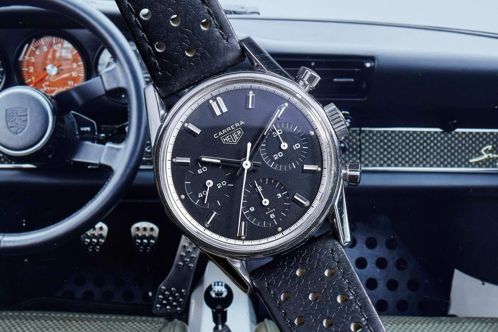 A Heuer Carrera 2447N watch with a black dial and perforated leather strap rests before a classic car dashboard, showcasing three sub-dials and silver hour markers. Detailed description coming soon!.
