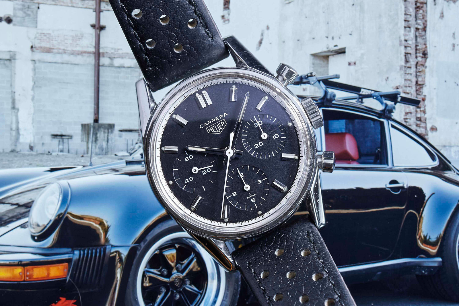 A close-up of a vintage Heuer Carrera 2447N wristwatch with a black leather strap and black dial, positioned in front of a classic black Porsche by a weathered, white brick building. Detailed Description Coming Soon!.