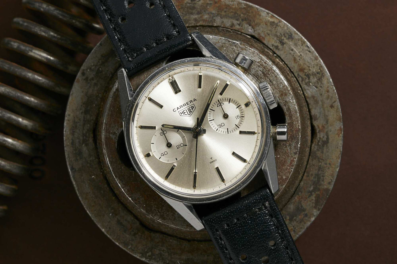 The Heuer Carrera by Heuer is a vintage racing chronograph with a silver dial, black leather strap, and two subdials, stylishly showcased on a large metal coil set against a brown background.