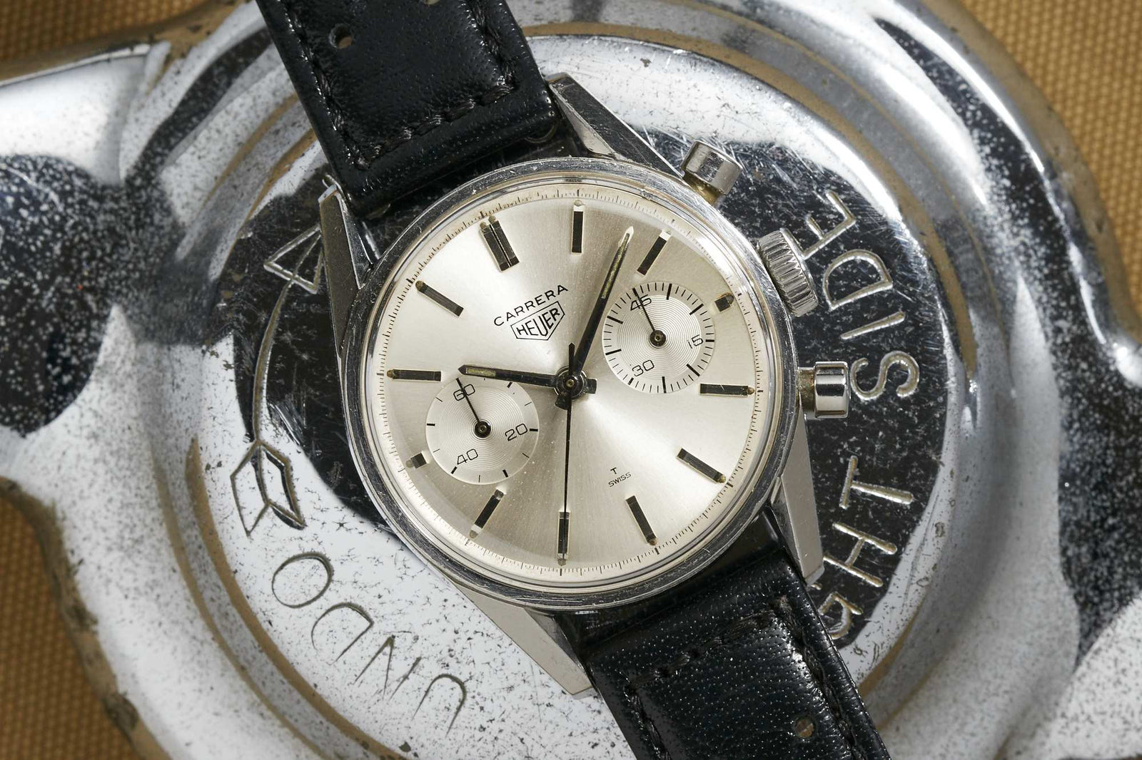 The Heuer Carrera by Heuer, inspired by Jack Heuer’s iconic racing chronograph, has a silver dial, black leather strap, and two subdials. It rests on a metallic object engraved “THE SIDE” and “DUNLOP.”.