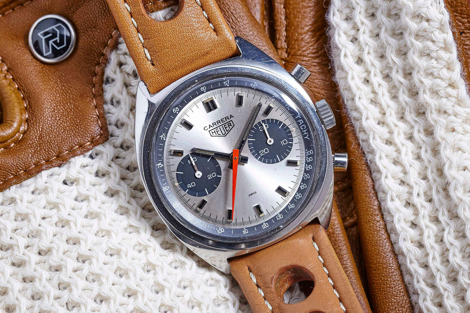 A Heuer Carrera Manual Wind by Heuer featuring a brown leather strap, silver dial with black subdials, and a red seconds hand is displayed on cream knit fabric atop a brown leather surface.