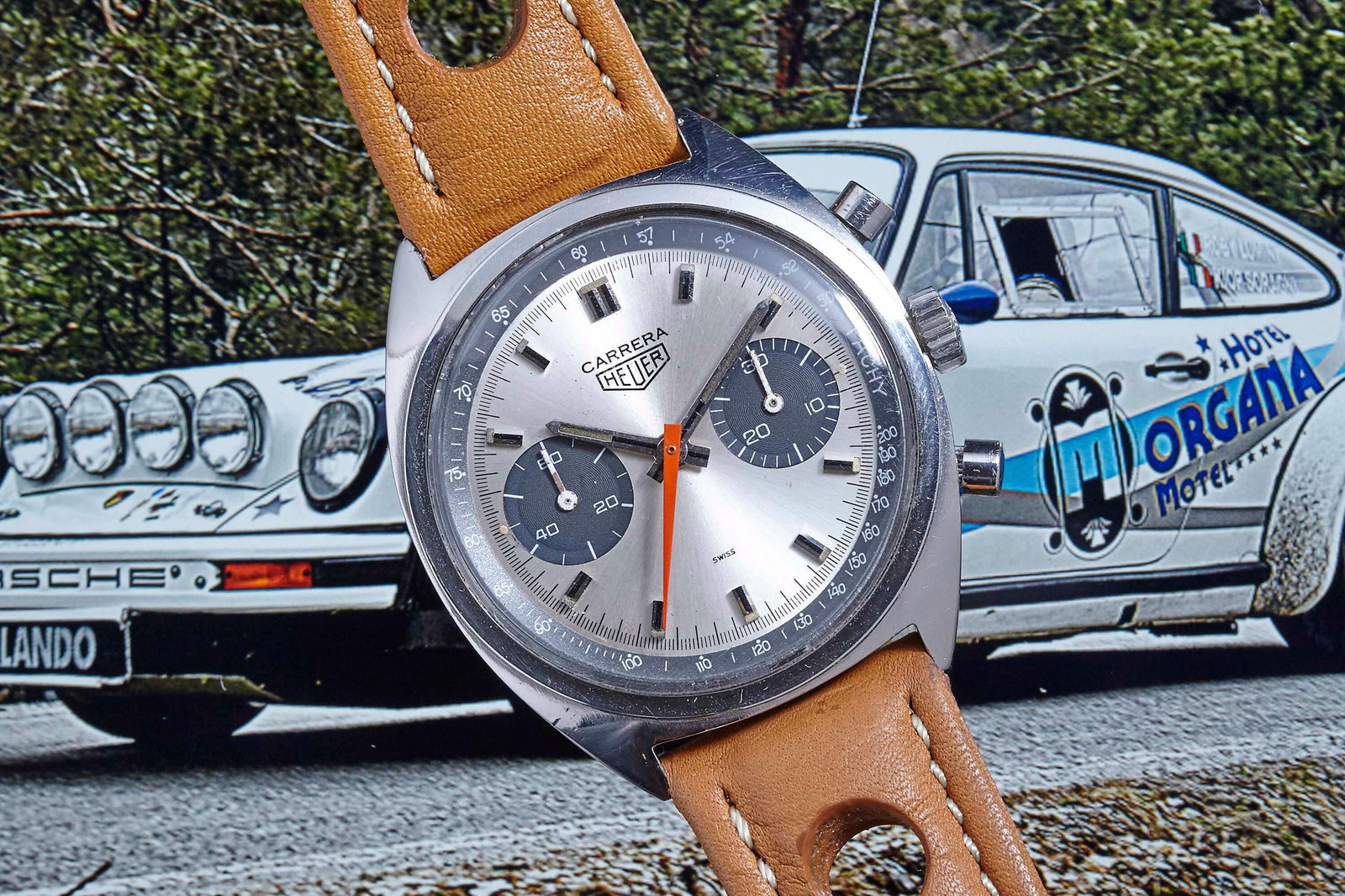 A close-up of the Heuer Carrera Manual Wind by Heuer with a tan leather strap is displayed in front of a classic white Porsche race car featuring racing decals and the word Morgana.
