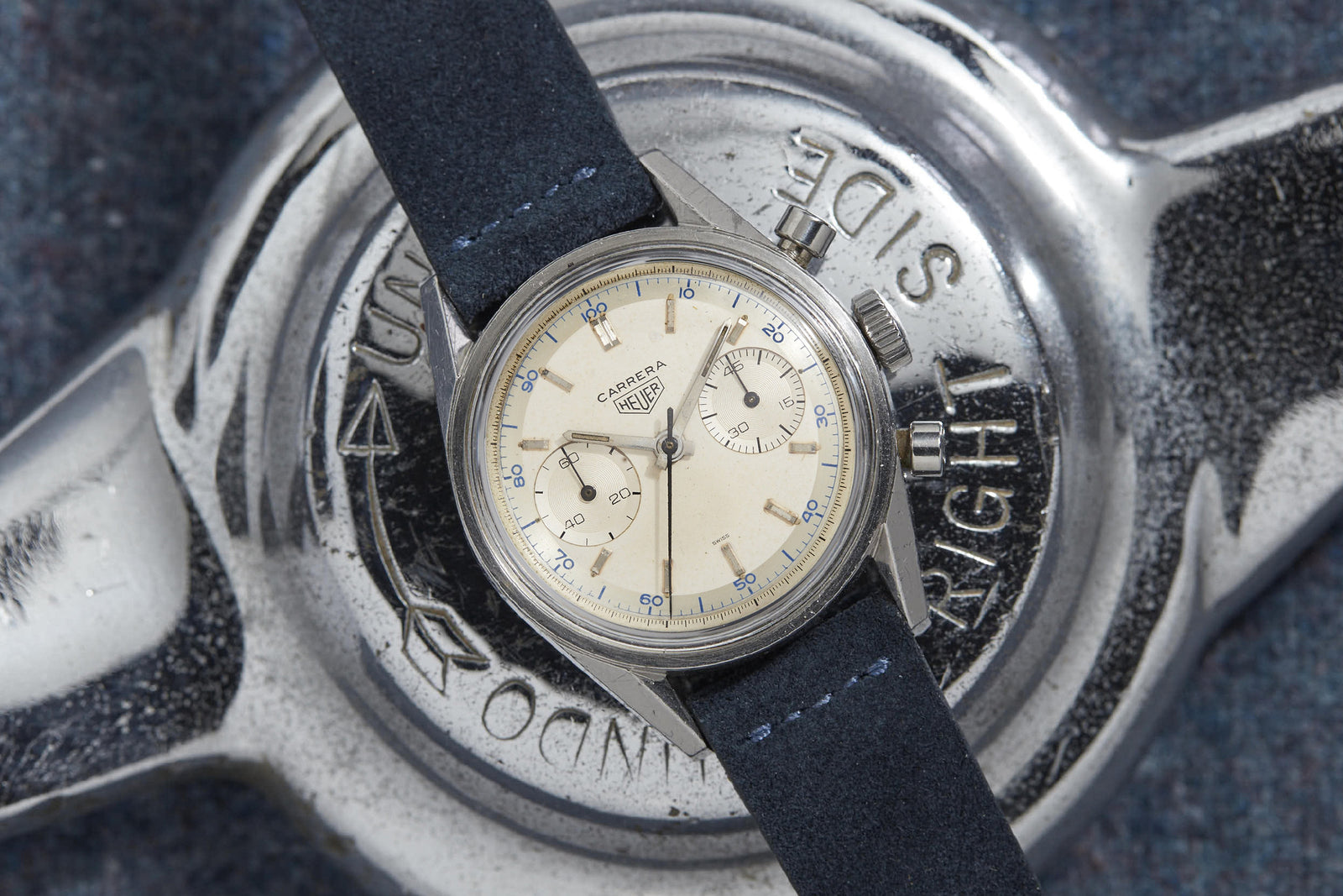 A vintage Heuer Carrera watch with a cream dial and blue subdials on a dark blue leather strap rests on an engraved metal object—celebrating Jack Heuer’s iconic tension ring innovation.