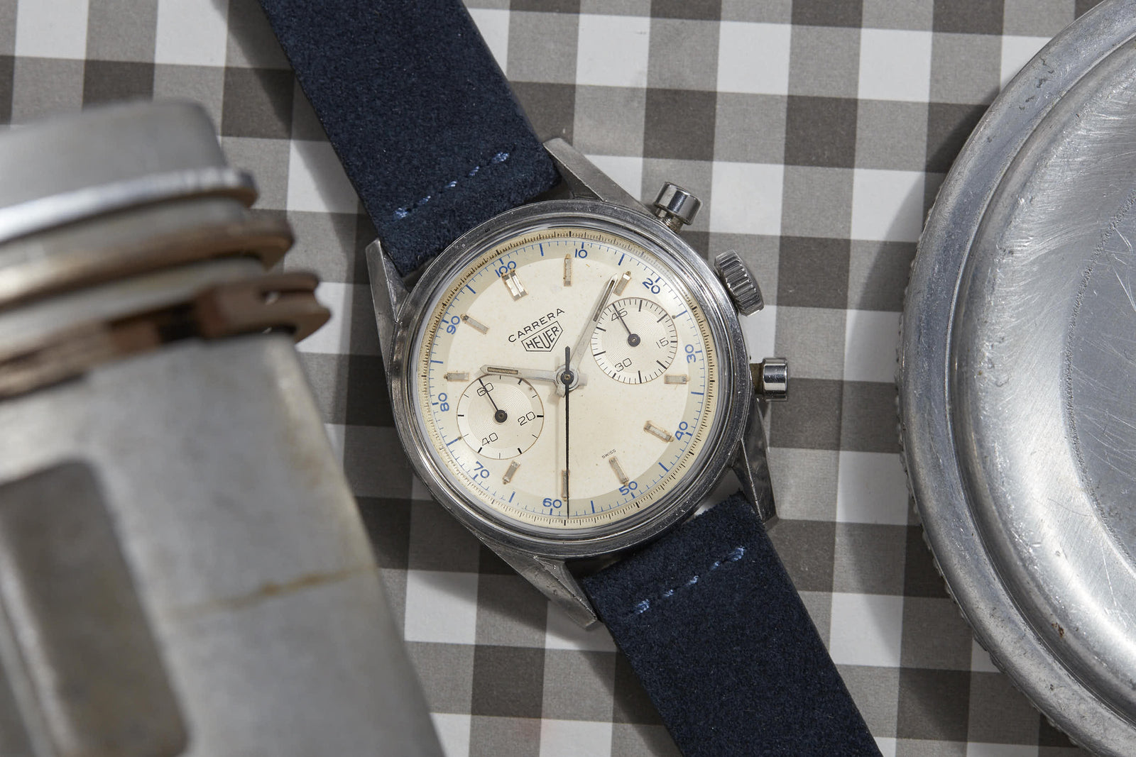 A vintage Heuer Carrera watch with a cream dial, two subdials, and a blue suede strap—honoring Jack Heuer’s tension ring innovation—rests on a gray-and-white checkered surface amid metallic objects.