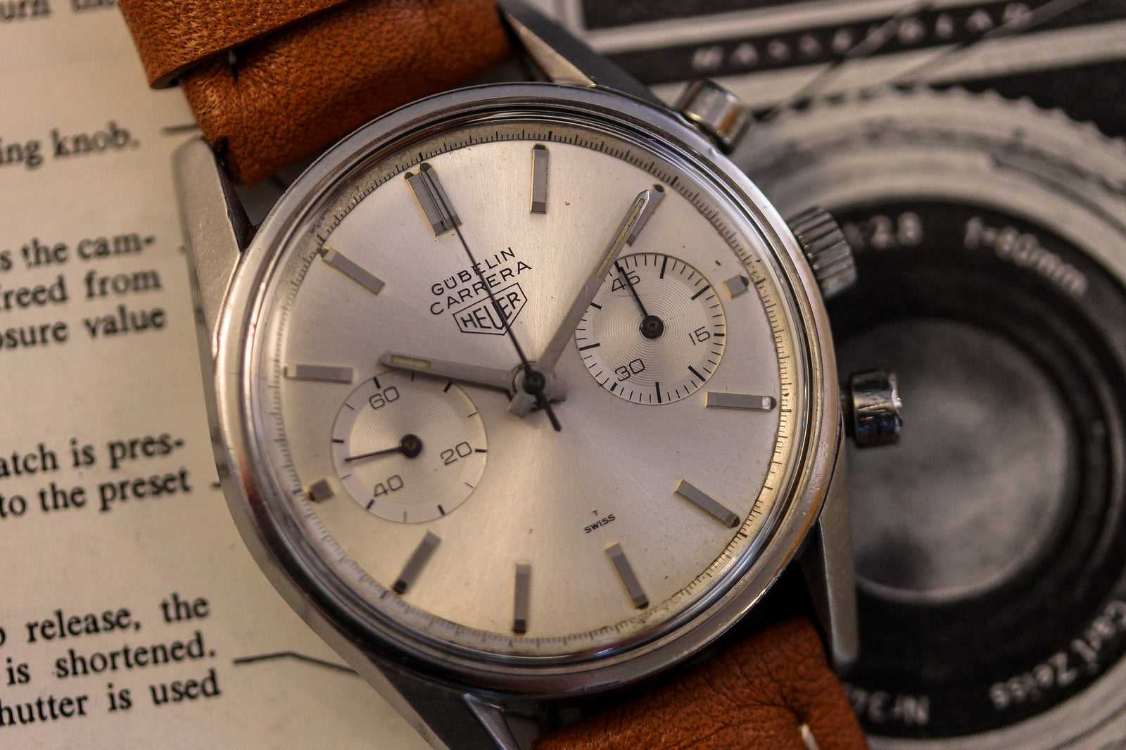 A Heuer Carrera Signed By Gübelin wristwatch with a silver dial and brown leather strap rests on an open book beside a black-and-white camera illustration.