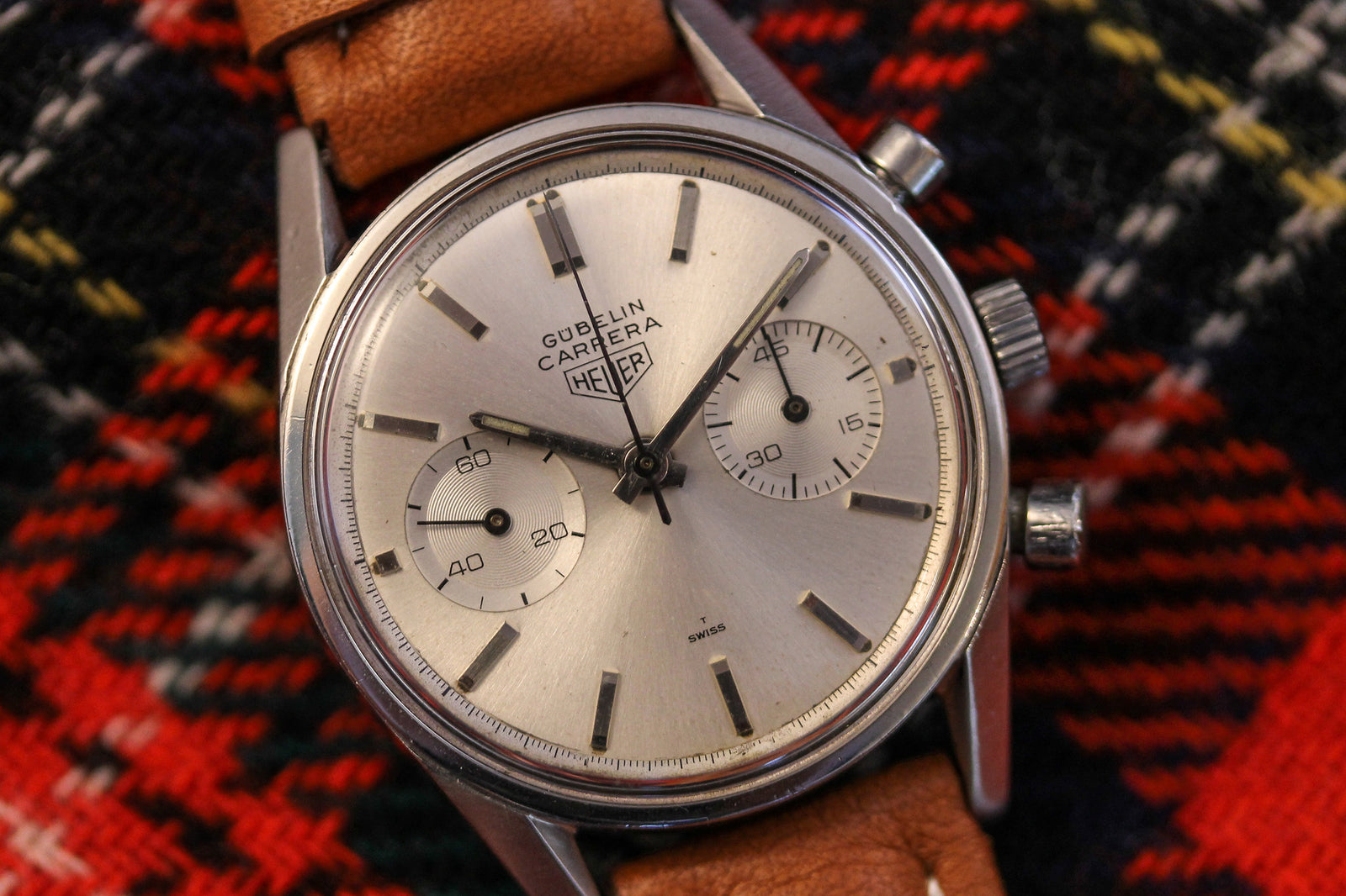 A Heuer Carrera Signed By Gübelin wristwatch with a tan leather strap, silver dial, and two subdials (Valjoux 92), is shown on a red and black plaid fabric background. The time displayed is approximately 8:16:57.