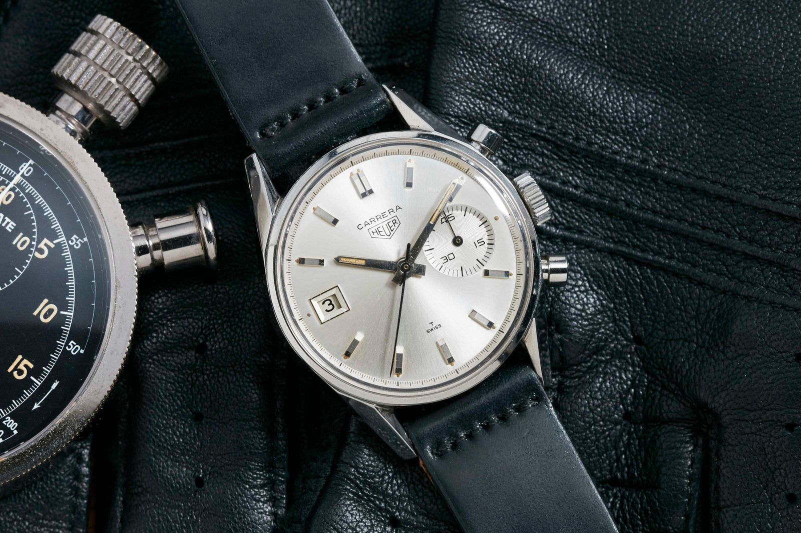 A Heuer Dato 3147S watch by Heuer, featuring a silver dial, date display, and elegant silver hour markers with a black leather strap. It rests on black leather gloves beside a vintage stopwatch.