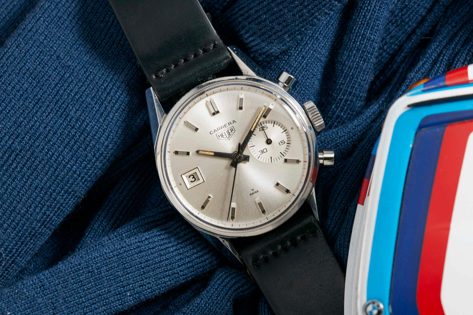 A Heuer Dato 3147S watch by Heuer, featuring a date display and sleek sub-dial, rests on blue fabric beside a white helmet decorated with red and blue stripes.