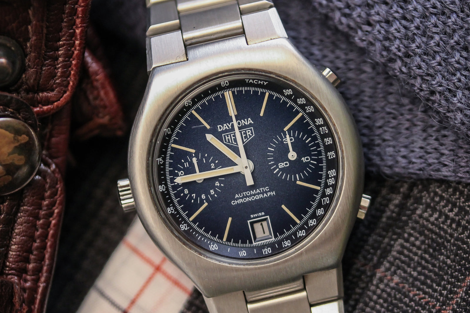 A close-up of a collector's watch: the Heuer Daytona Chronograph - NOS by Heuer, featuring a black dial, three subdials, and a date window, elegantly framed by textured clothing and leather fabric.