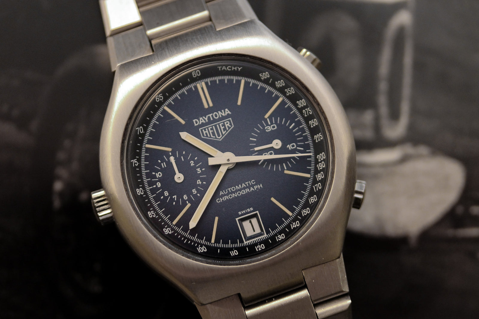 Close-up of a Heuer Daytona Chronograph - NOS features a blue dial with three subdials, date window, detailed white indices, tachymeter markings, and a stainless steel bracelet—ideal for Heuer collectors and steel chronograph enthusiasts.