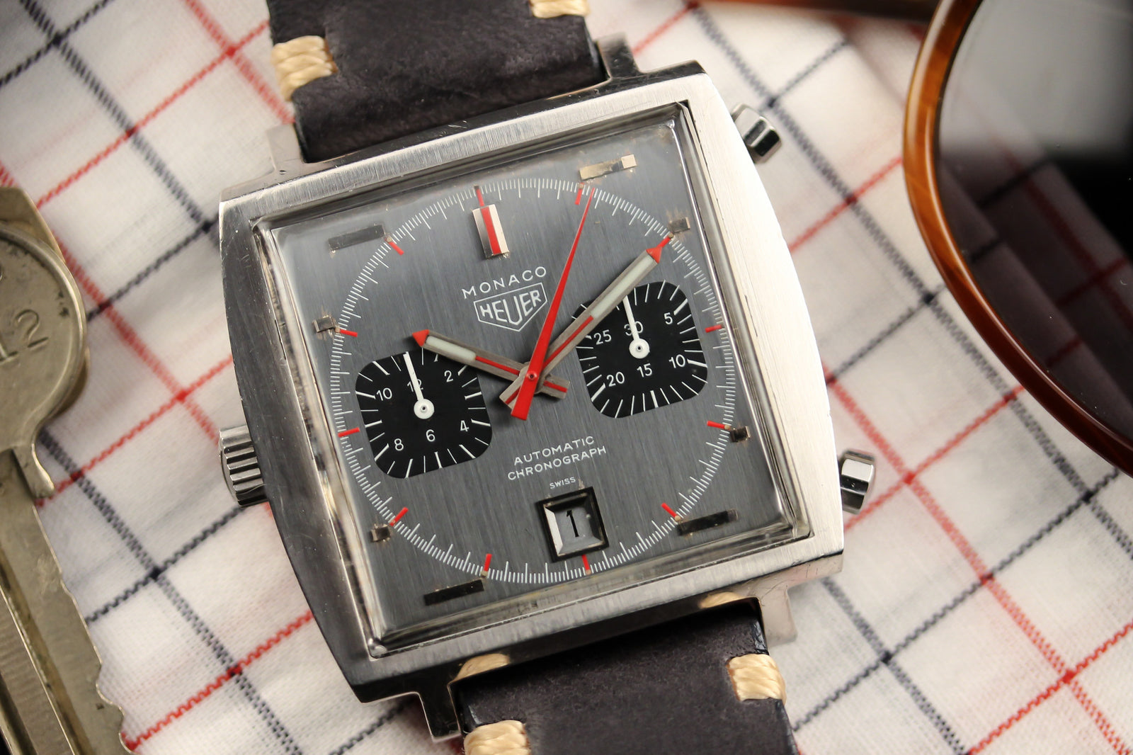 A Heuer Monaco 1133G, a square chronograph with a gray dial, black subdials, red hands, and black leather strap from Heuer, rests on plaid cloth beside a key and part of sunglasses.