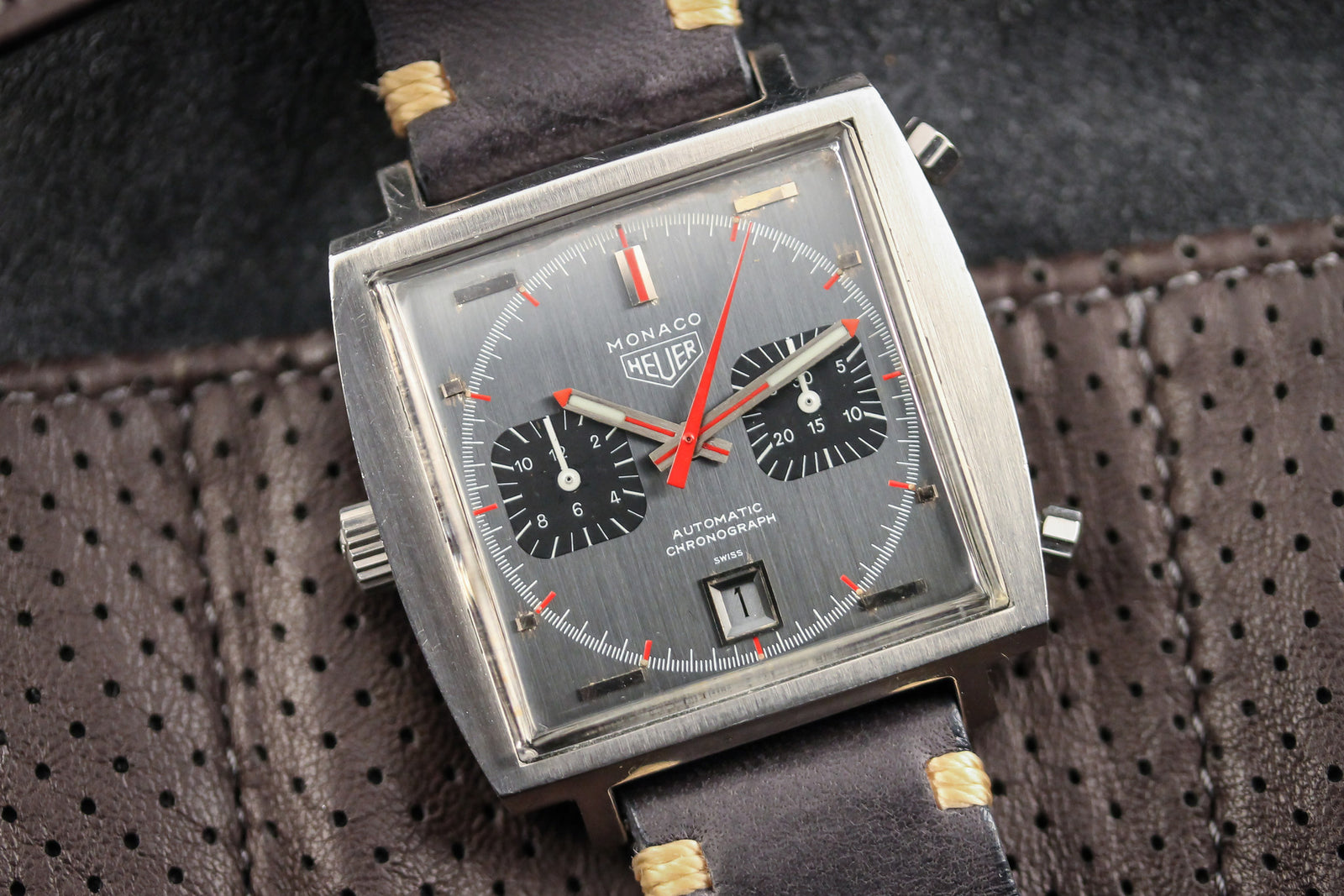 The Heuer Monaco 1133G features a square case, black dial with two subdials, red hands, and a date at 6 o'clock, paired with a dark leather strap with beige stitching, displayed on brown perforated leather.