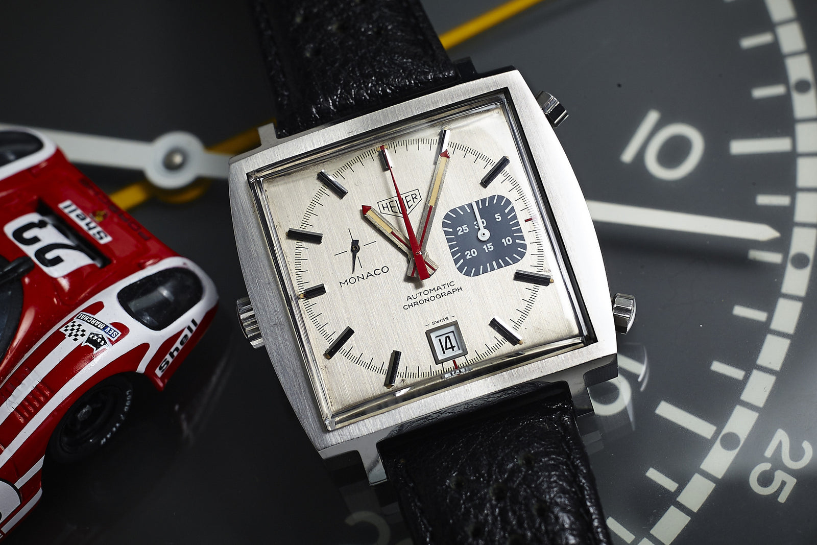 A Heuer Monaco 1533G chronograph wristwatch with a black leather strap rests on a clock face beside a red and white toy race car. This classic Heuer piece features date display and a blue subdial, reminiscent of Steve McQueen’s iconic style.
